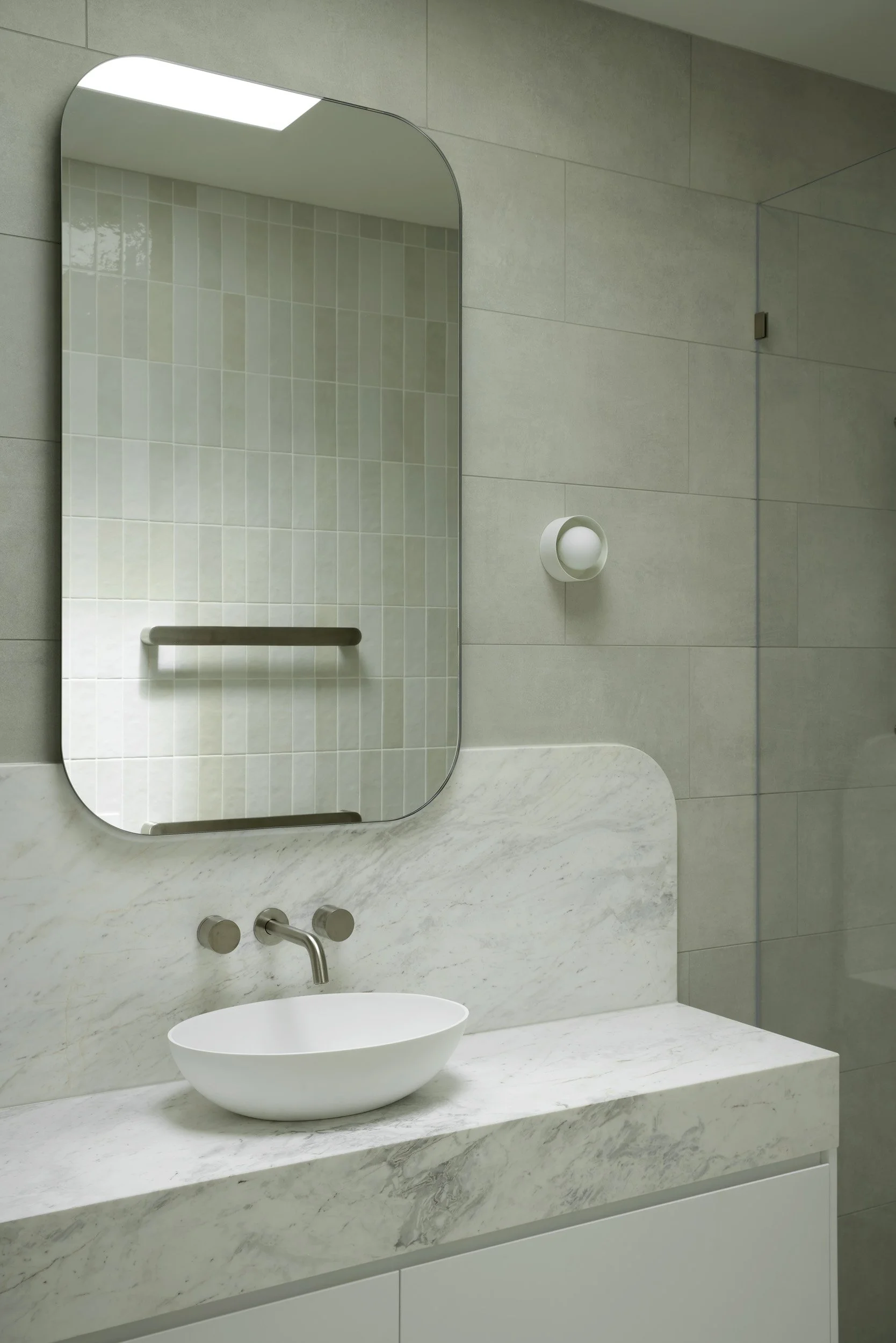 Modern bathroom with a gray tiled wall, a large rectangular mirror, a white marble sink countertop, a vessel sink, and a wall-mounted faucet. There is a small wall light next to the mirror.