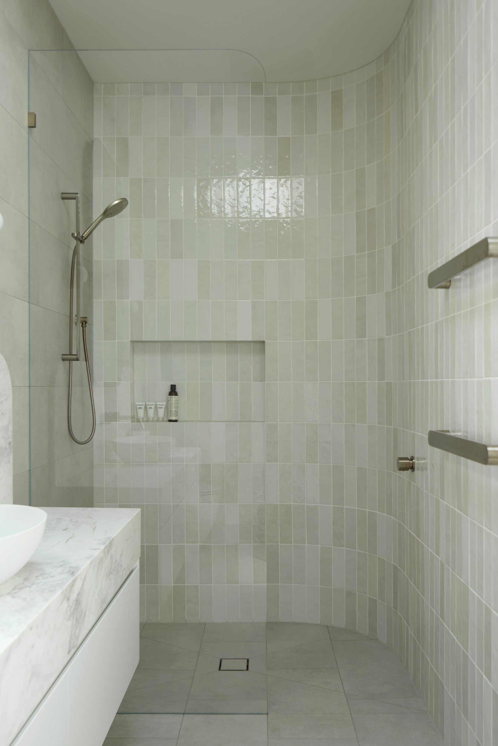 Empty modern shower with beige tiles, a built-in niche with toiletries, wall-mounted showerhead, and a marble-topped vanity.