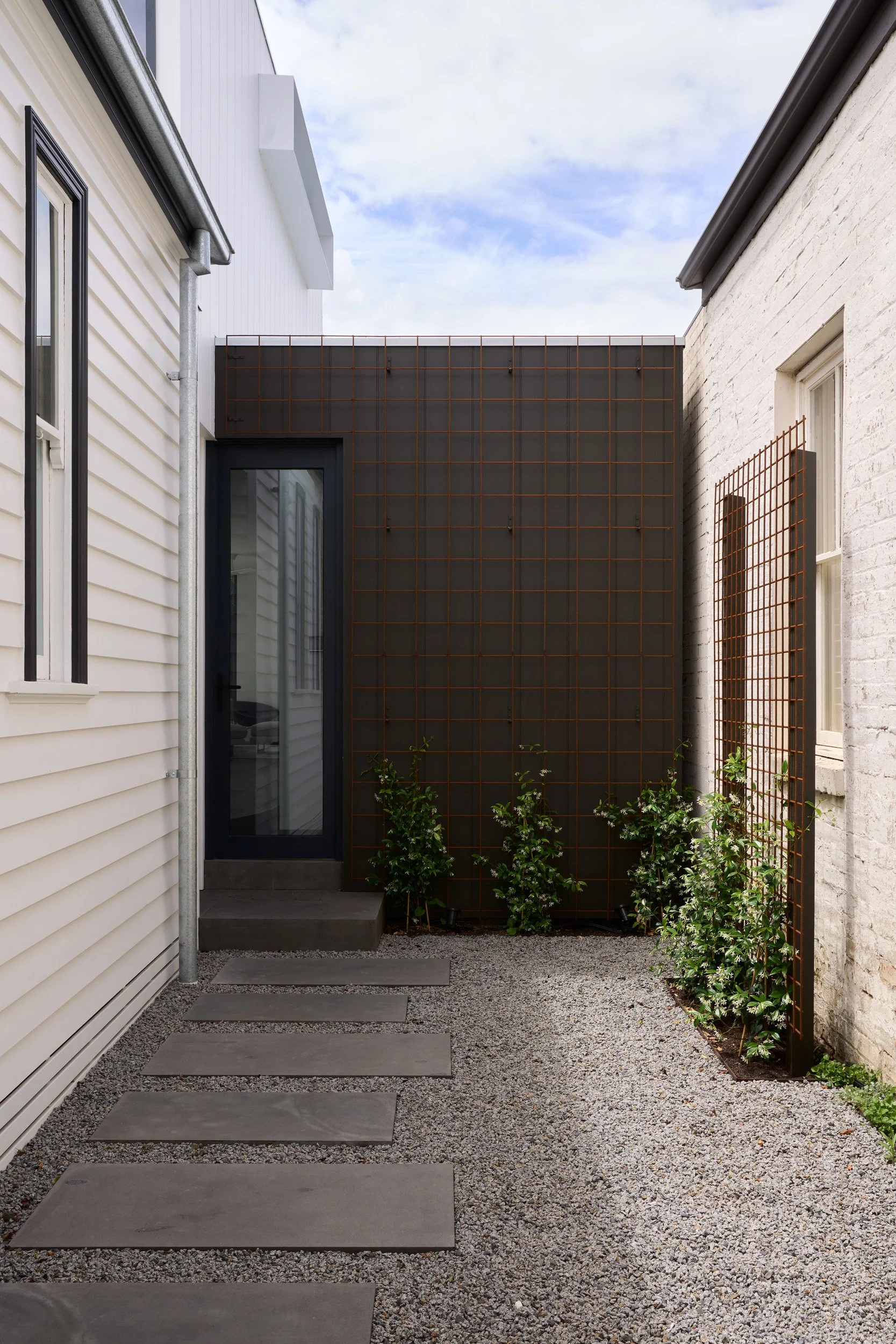 Small outdoor space with gray concrete stepping stones, gravel ground, a black door with glass, white and gray houses on either side, and some green plants along the base of a rust-colored metal trellis.
