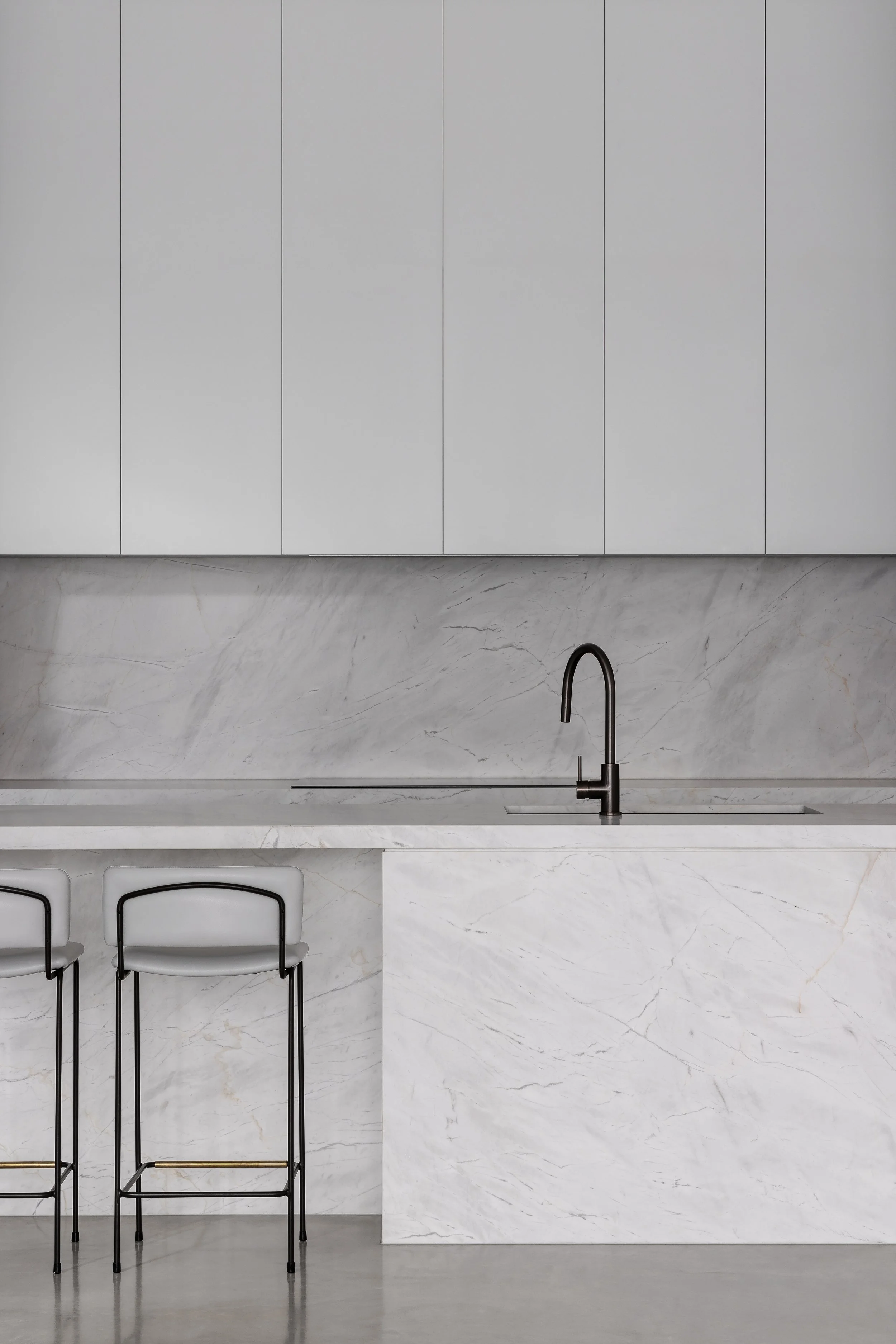 Modern minimalist kitchen with white marble island, backsplash, and sleek white cabinets, featuring a black faucet and two white bar stools.