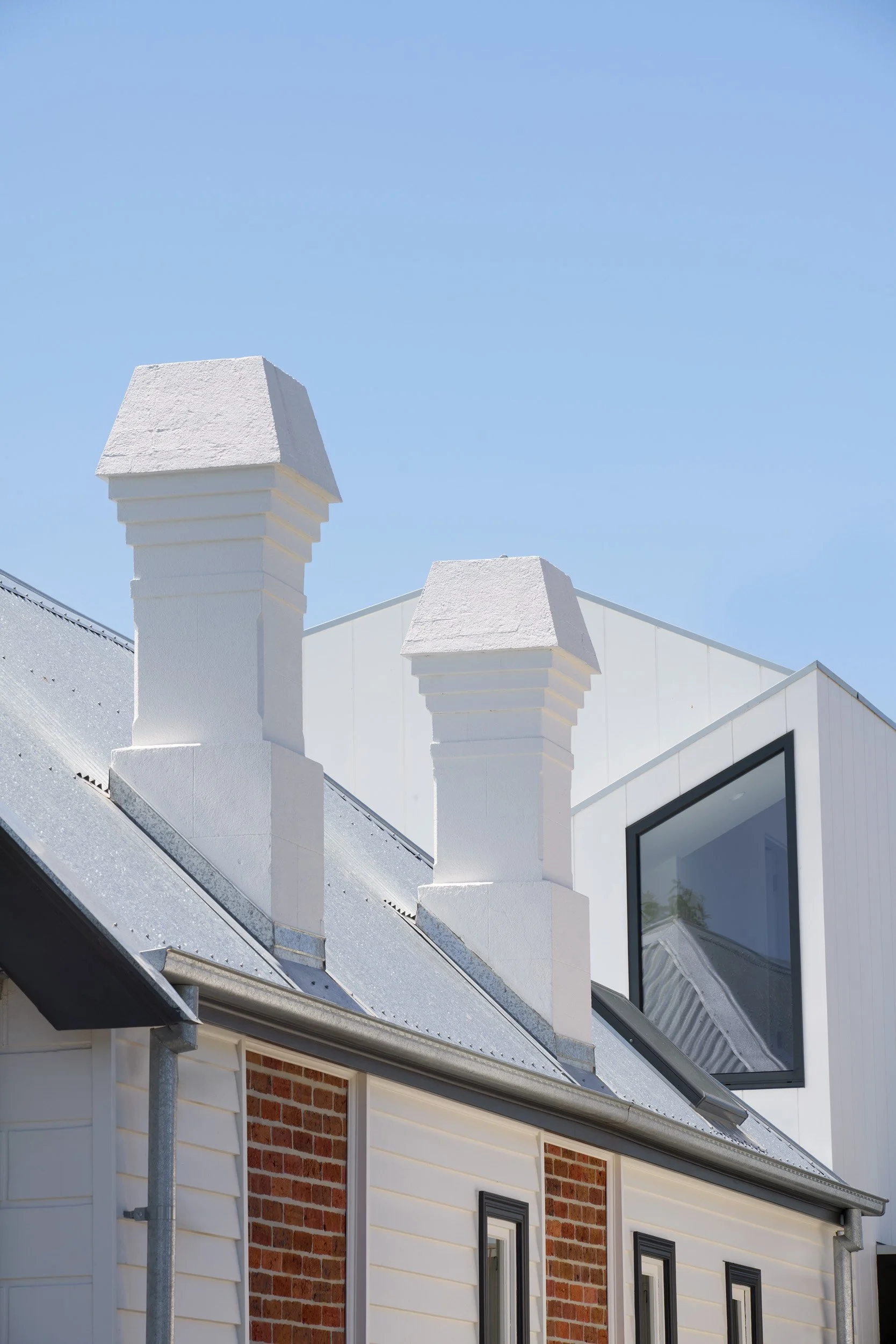 Close-up of two white chimneys on a modern house with a sloped metal roof, brick and siding exterior, and a large window, under a clear blue sky.