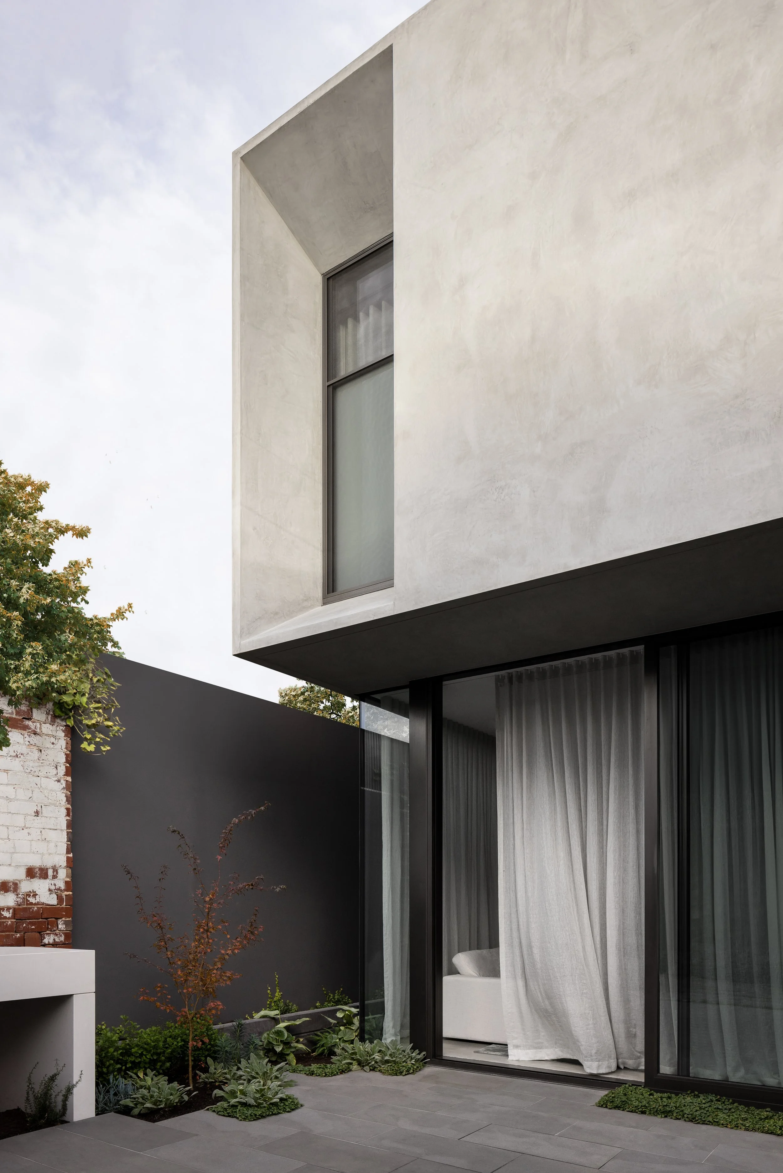 Modern house with white and black exterior, large windows with curtains, a small garden with plants and a tree, and a patio area.