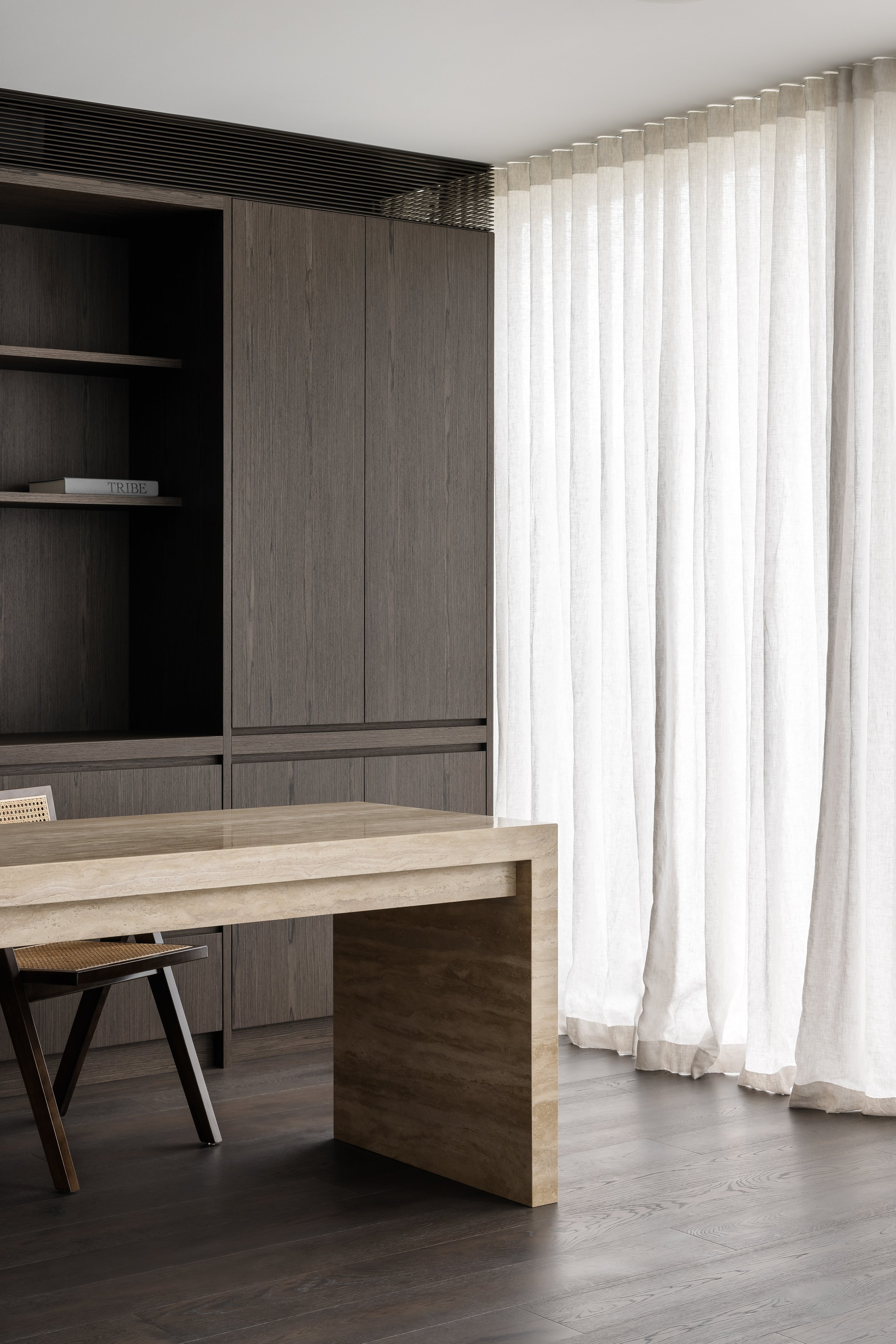 Modern office space with a light-colored stone desk, dark wood chair, gray built-in shelving, and long white curtains covering a window.