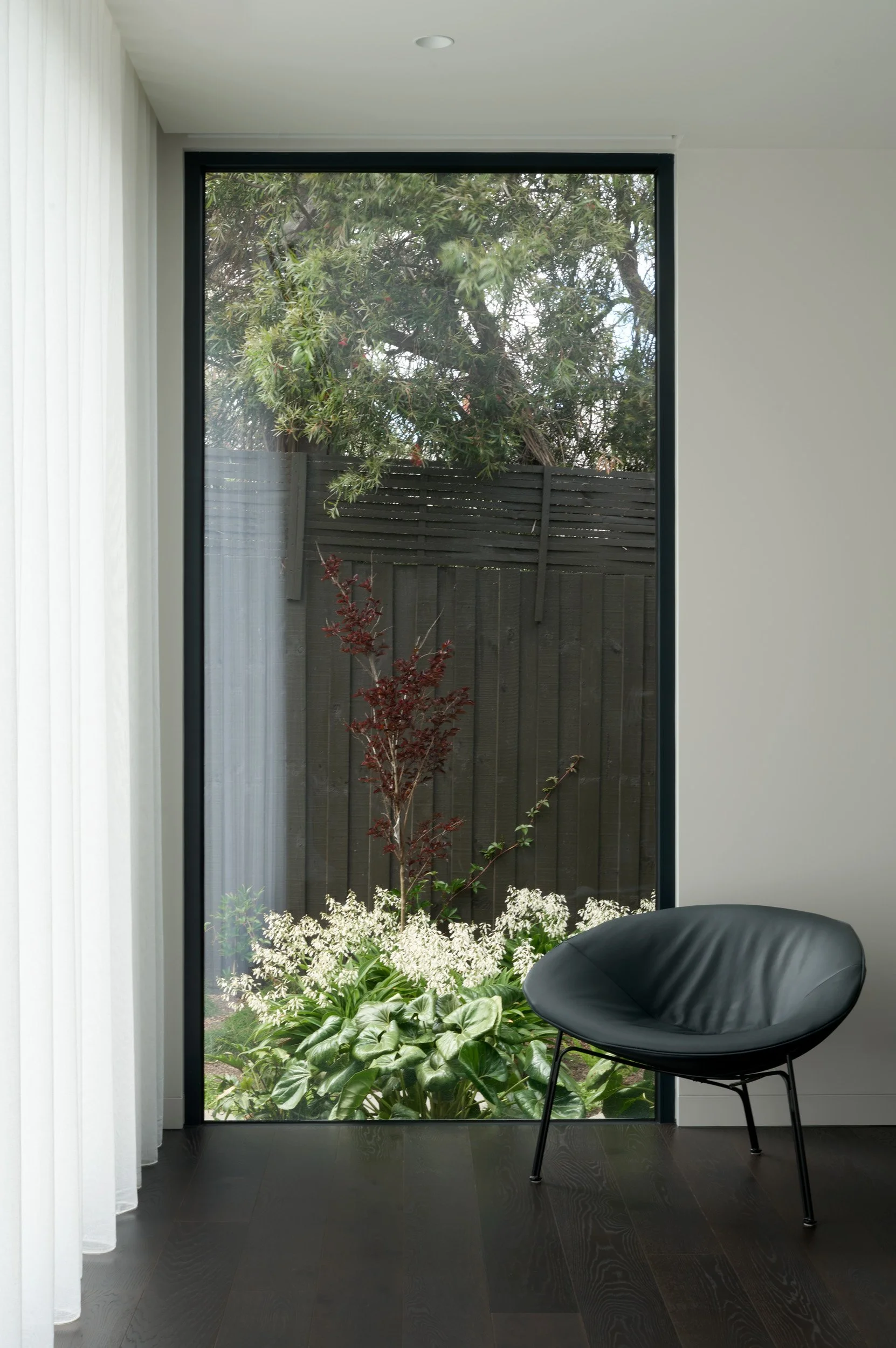 Modern room with a black leather chair beside a large floor-to-ceiling window, view of a backyard with plants, trees, and a wooden fence.