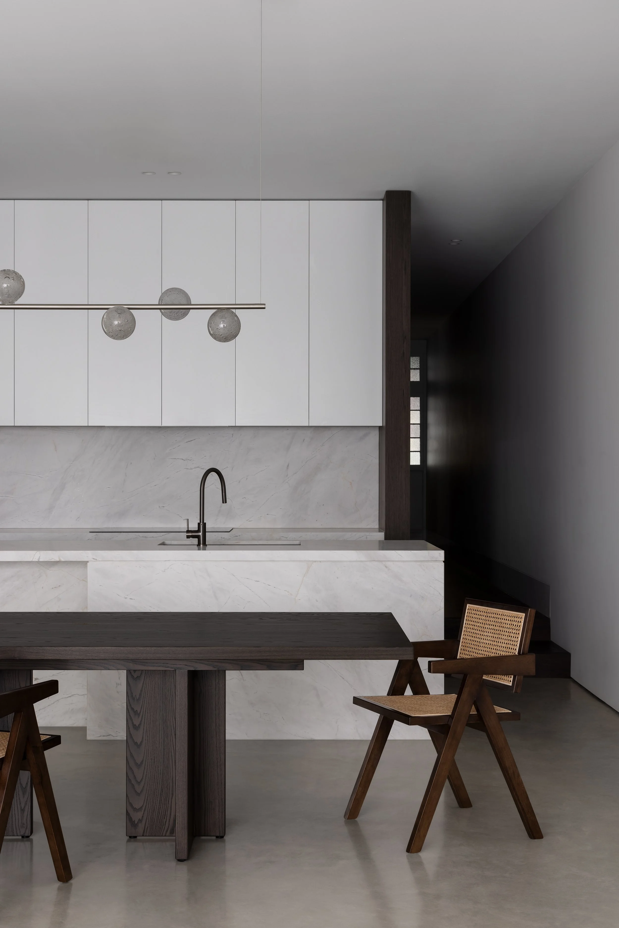 Minimalist kitchen with white cabinetry, marble backsplash and countertop, black faucet, wooden dining table, and two wooden chairs with cane seats.