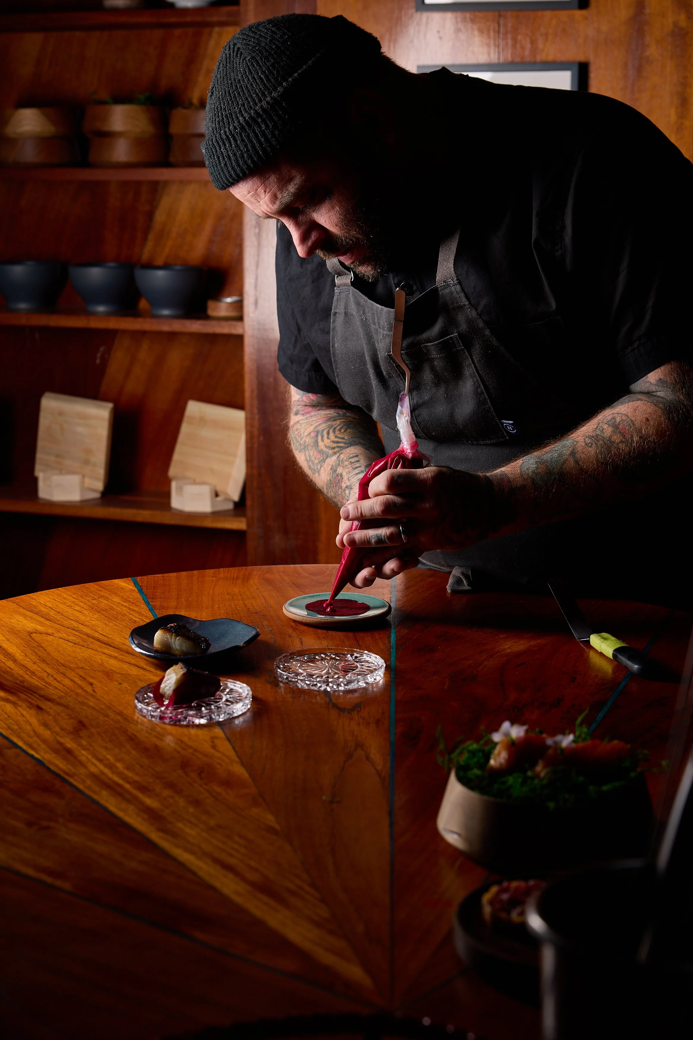 A chef, wearing a black beanie and black apron, prepares to plate a dish in a restaurant kitchen with wood-paneled walls and shelves holding black bowls.