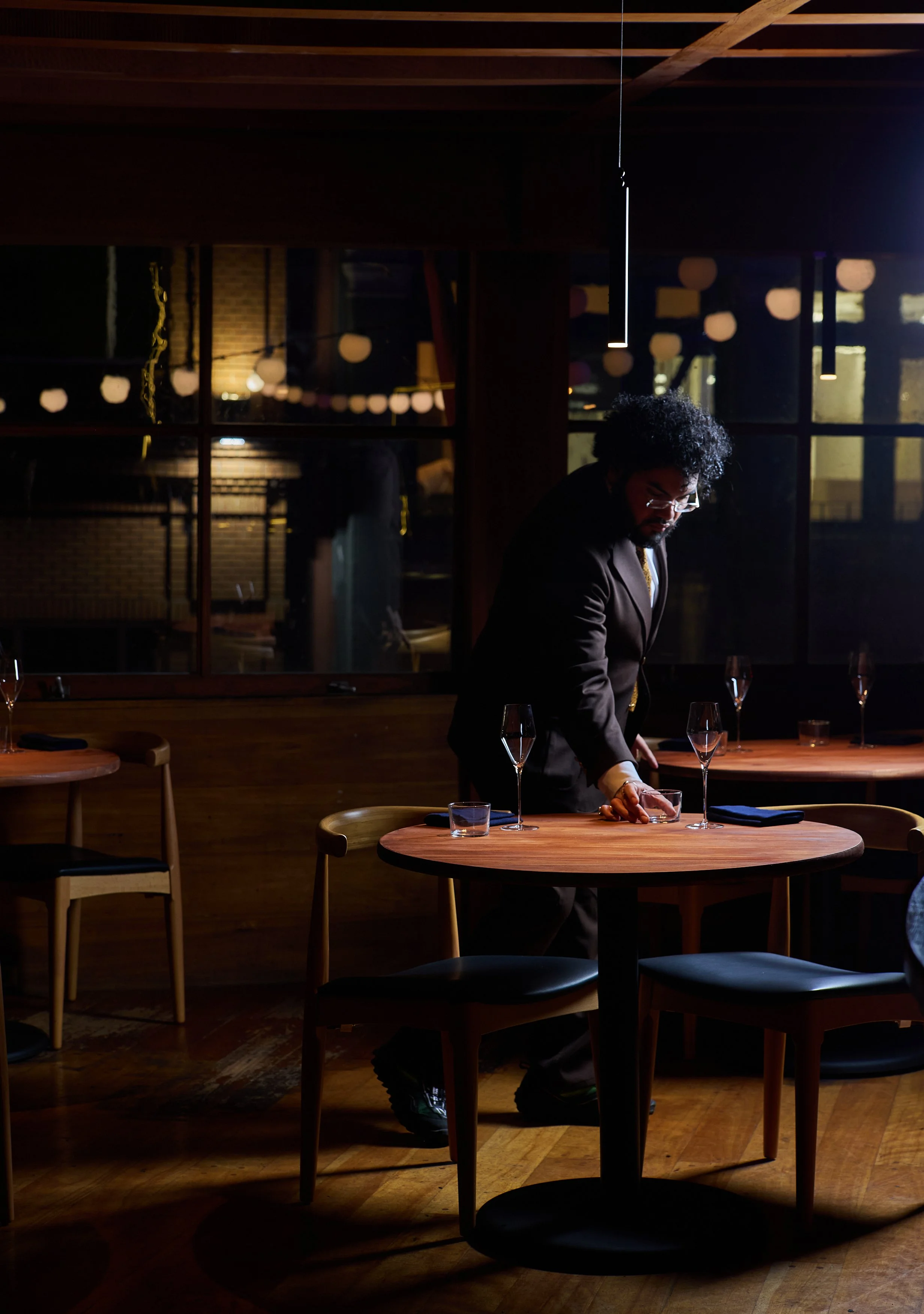 A man with curly hair and glasses, dressed in a suit, arranging wine glasses on a wooden table in The Lore's dimly lit salon with a window showing decorations outside.