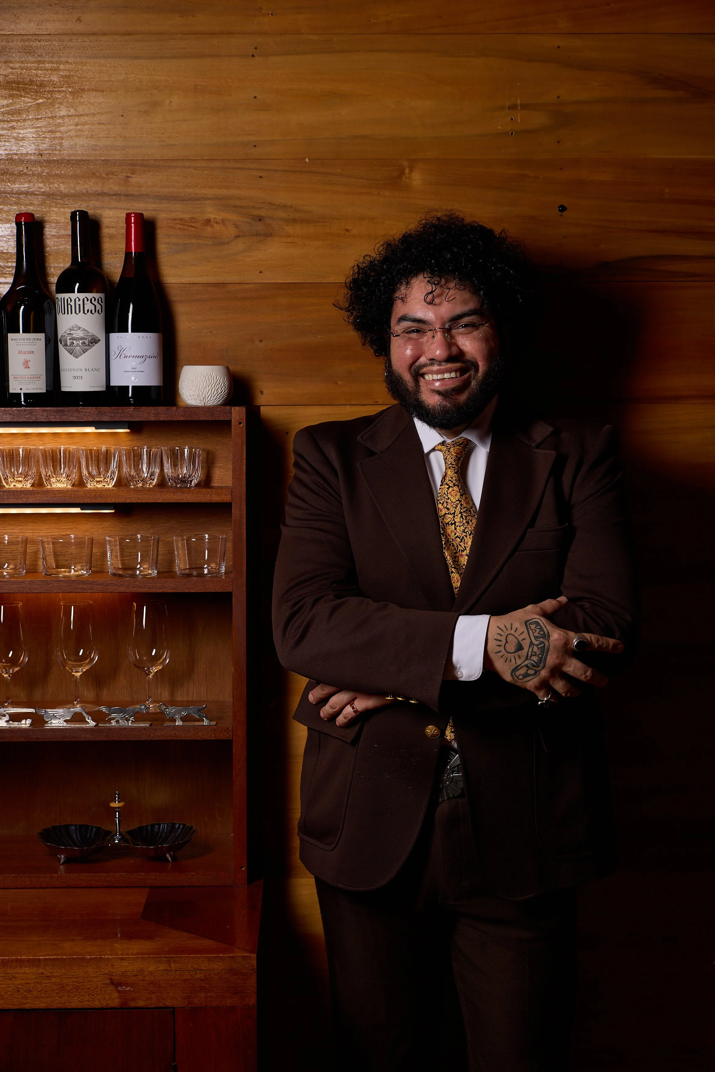 A man with curly black hair and glasses, wearing a dark suit and patterned tie, standing against a wooden wall with a wine rack and glasses behind him, smiling at the camera.