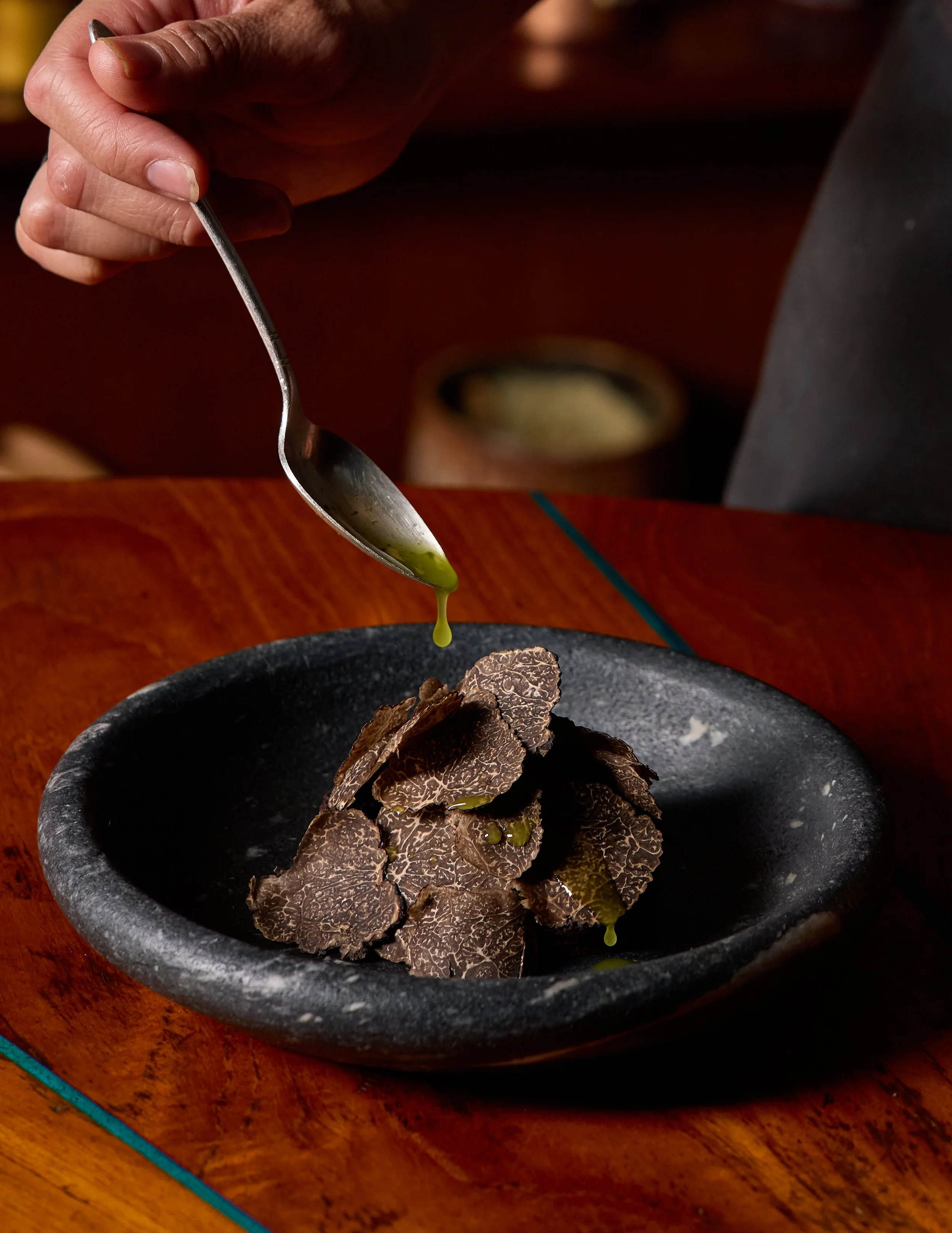 Plating of thinly sliced black truffles topped with olive oil on a black bowl, placed on a wooden surface. Example of a dish at The Lore.
