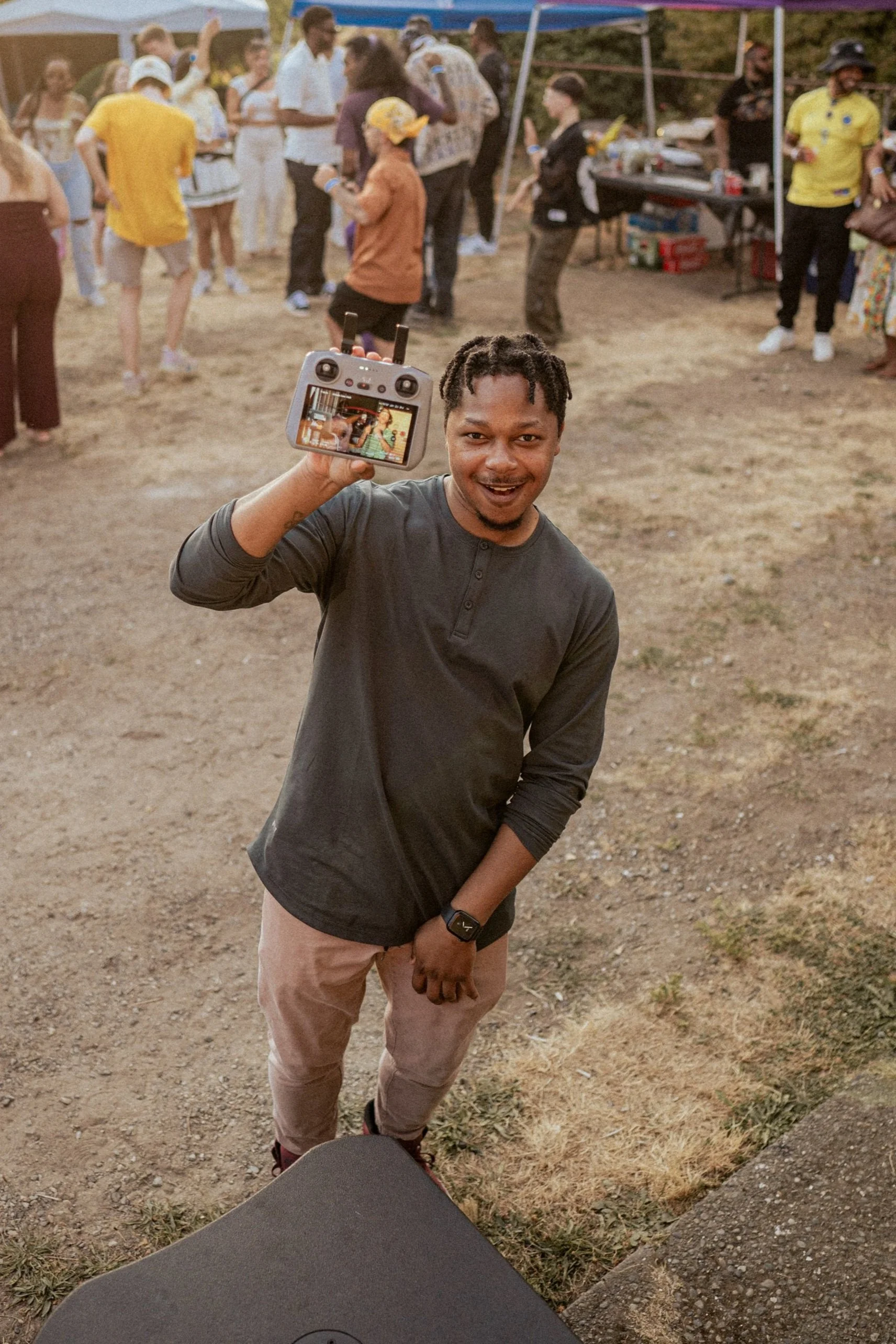 A man smiling and holding a remote control device while standing at an outdoor event with a crowd of people in the background.