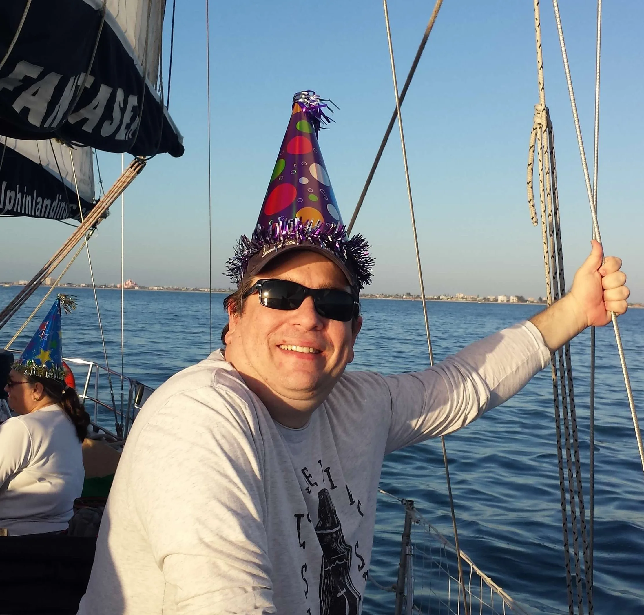 Warren wearing sunglasses and a colorful birthday party hat on a boat, smiling, with water and a distant shoreline in the background.