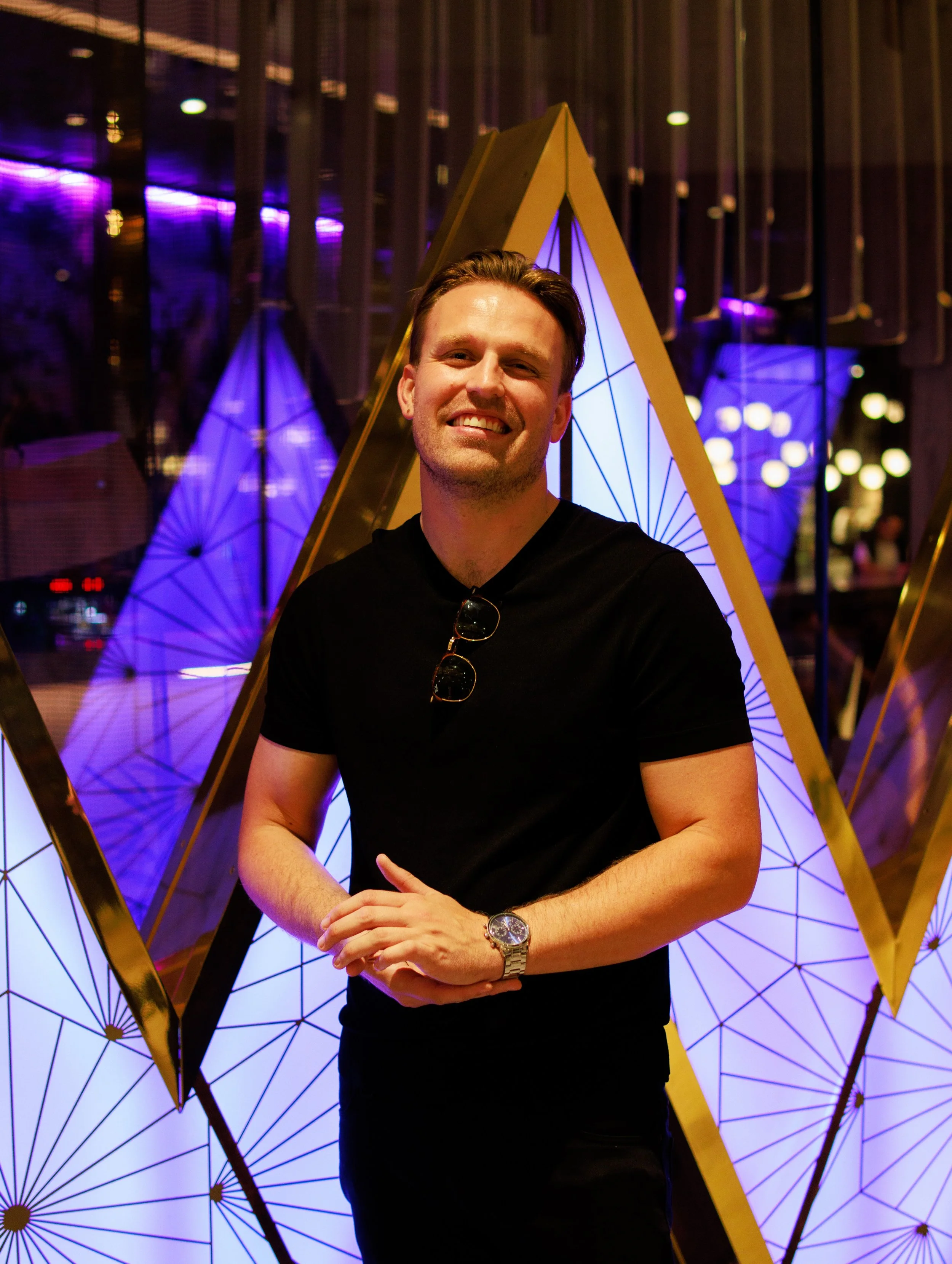 A smiling man in a black t-shirt standing in front of illuminated geometric art installation at night.