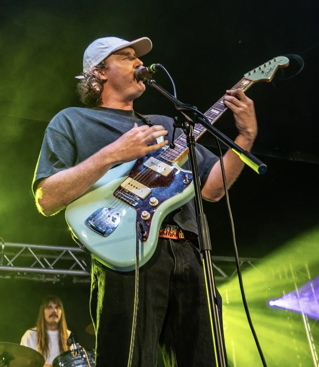 A male musician singing into a microphone while playing an electric guitar on stage with colorful lighting.