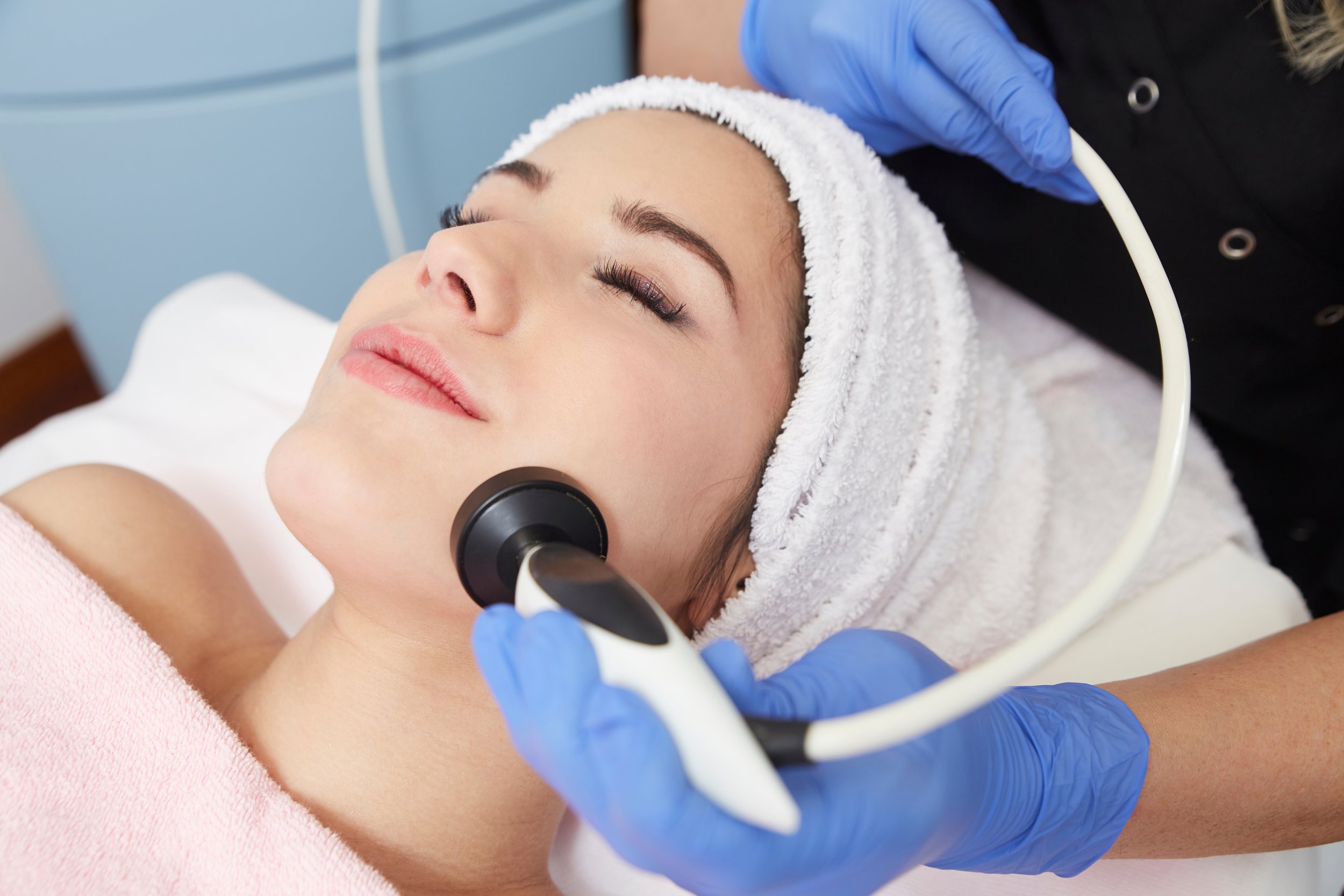 Woman receiving a facial treatment using a handheld device at a spa or dermatologist's office, with a towel on her head and a pink towel around her shoulders.