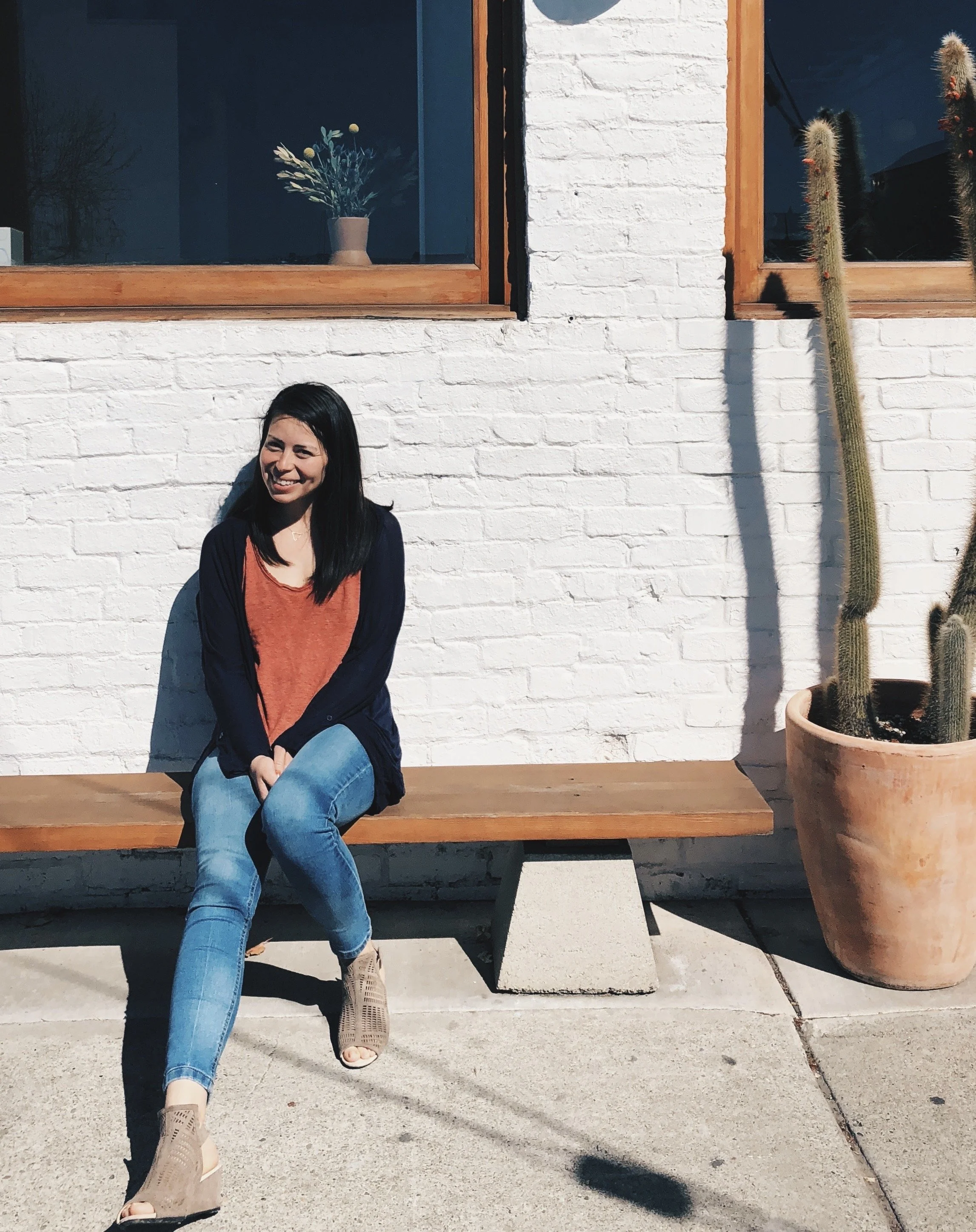 A woman sitting on a wooden bench outside a building with a white brick wall, a large potted cactus, and a window with a flower vase on the sill.