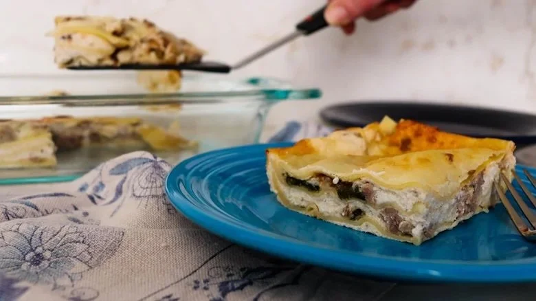 A slice of white lasagna on a blue plate, with additional slices in a glass baking dish in the background. A person is lifting a portion of the lasagna with a spatula.