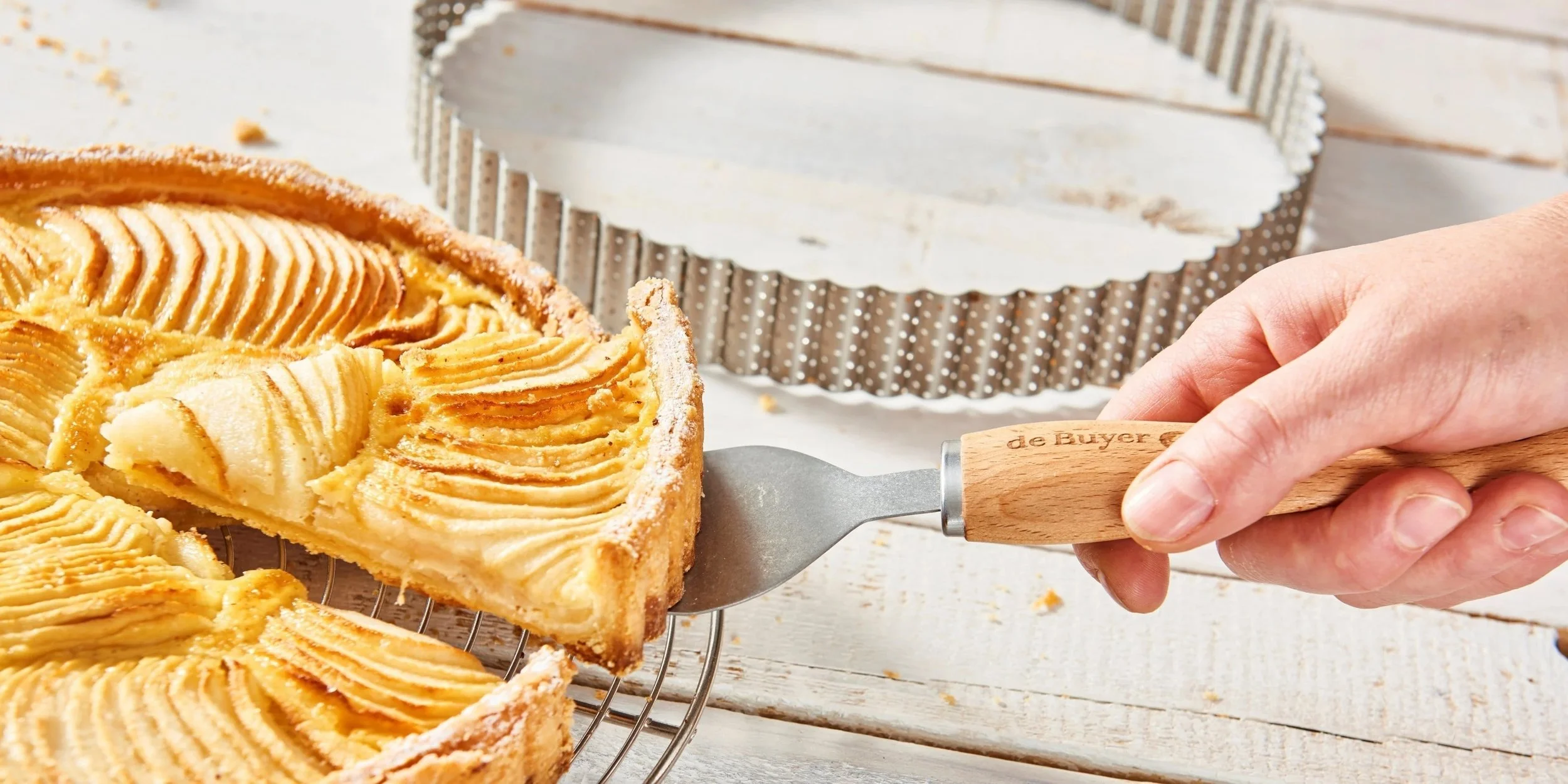 A person is lifting a slice of apple tart with a spatula, showing the layered apple slices on top, with a partially empty tart in the background.