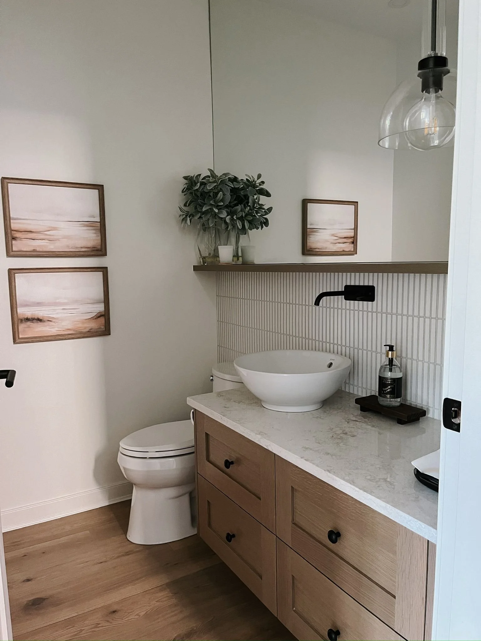 Bathroom with wooden vanity, vessel sink, black faucet, toilet, wall mirror, framed artwork, potted plant, pendant light, and soap dispenser.