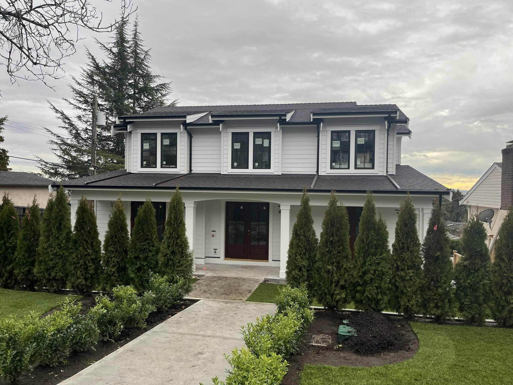 A two-story house with white siding, black roof, and multiple windows, surrounded by a well-maintained lawn with a concrete walkway leading to the front door, and tall green shrubs lining the walkway.