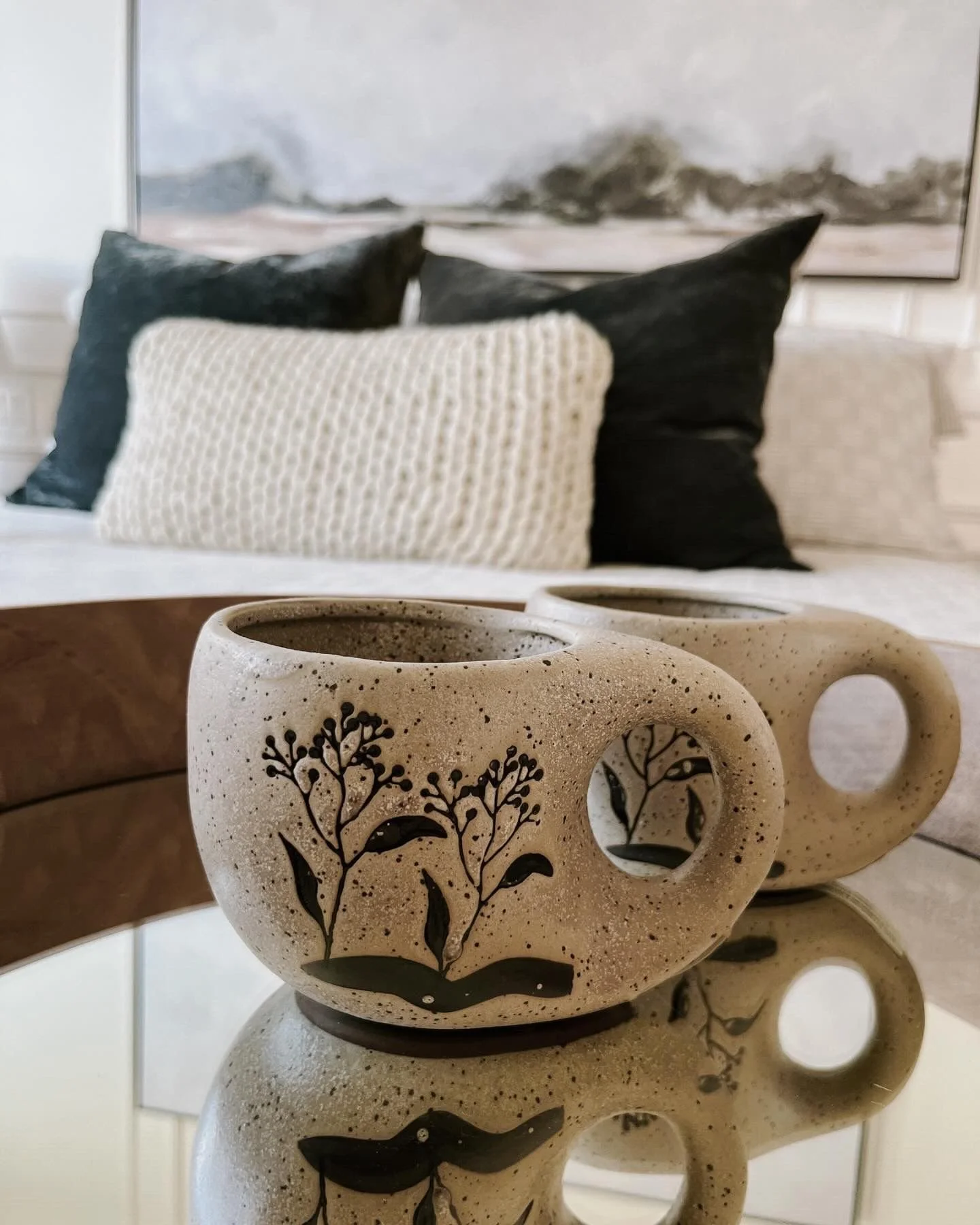 Two ceramic mugs with black floral designs, placed on a reflective glass surface in front of a beige sofa with black and white pillows, and a framed abstract black and white artwork on the wall.