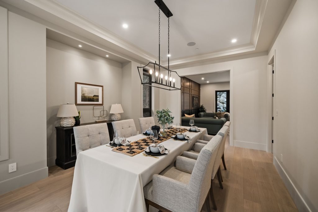 Modern dining room with a rectangular table set for dinner, surrounded by upholstered chairs, modern chandelier overhead, and a sitting area with sofas in the background.