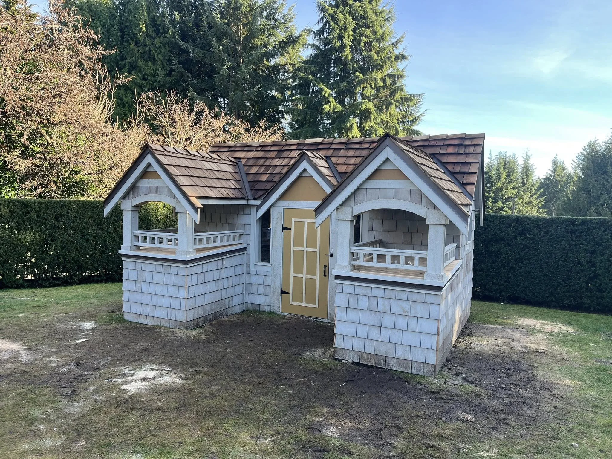 Miniature house in a yard with a shingled roof, small porches, and a decorative door, surrounded by grass and trees.