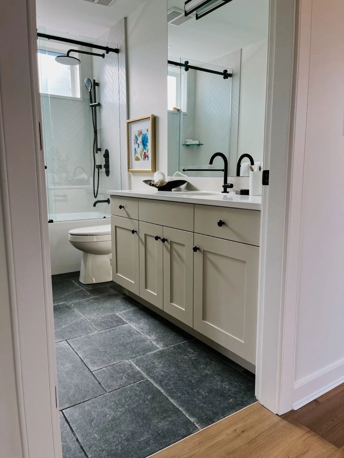 A bathroom with a white vanity, black fixtures, a mirror, a framed colorful art piece, a black bowl with a towel, and a shower with glass doors, black hardware, and a window.