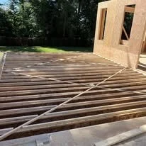 Construction site with wooden floor framing being built beside a brick building, with a hose on the ground and trees in the background.