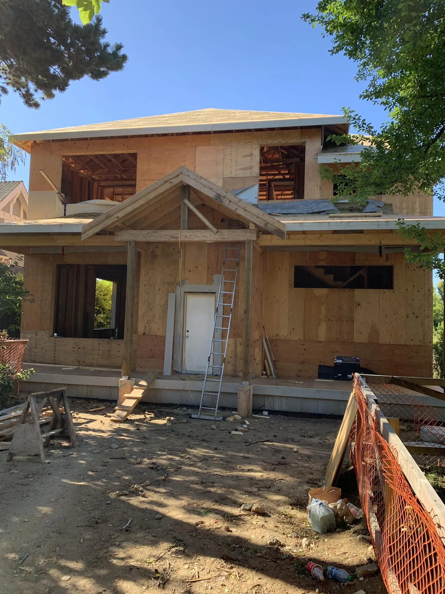 A two-story house under construction with plywood walls, a ladder leaning against the front, and an orange safety fence around the dirt yard.