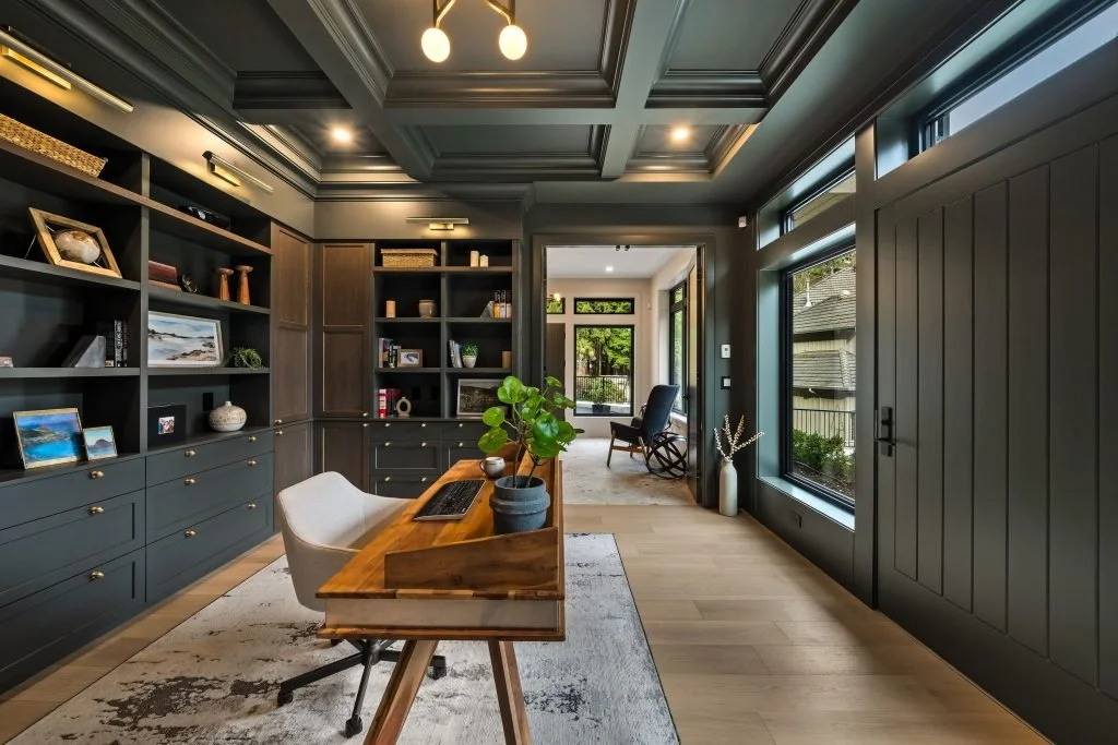 A modern home office with dark gray walls and built-in bookshelves, a wooden desk with a potted plant, an office chair, and large windows letting in natural light.