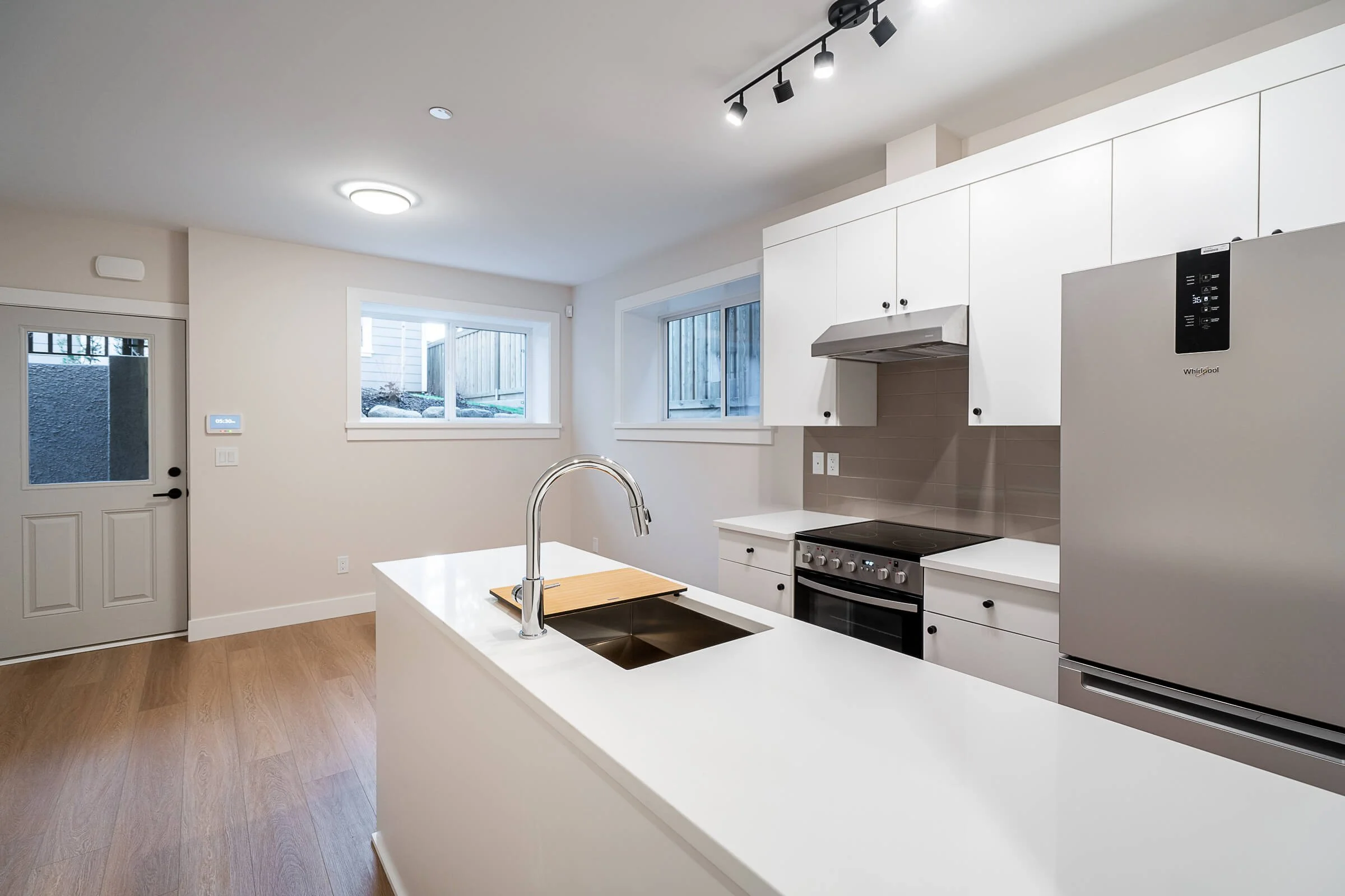 Modern kitchen with white cabinets, stainless steel refrigerator, black stove, and wooden flooring, with two windows and ceiling track lighting.