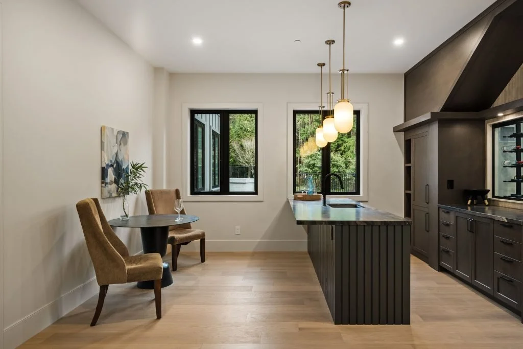 Modern kitchen with a small dining area, black cabinetry, pendant lighting, and large windows showing greenery outside.