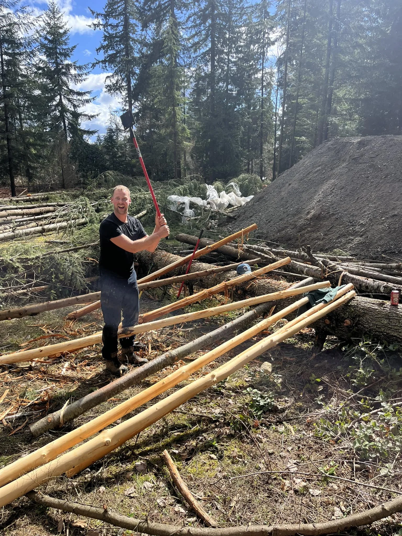 A man standing in a forest clearing, smiling and holding a red tool, surrounded by cut tree logs and poles, with a large pile of dirt in the background and tall trees and blue sky overhead.