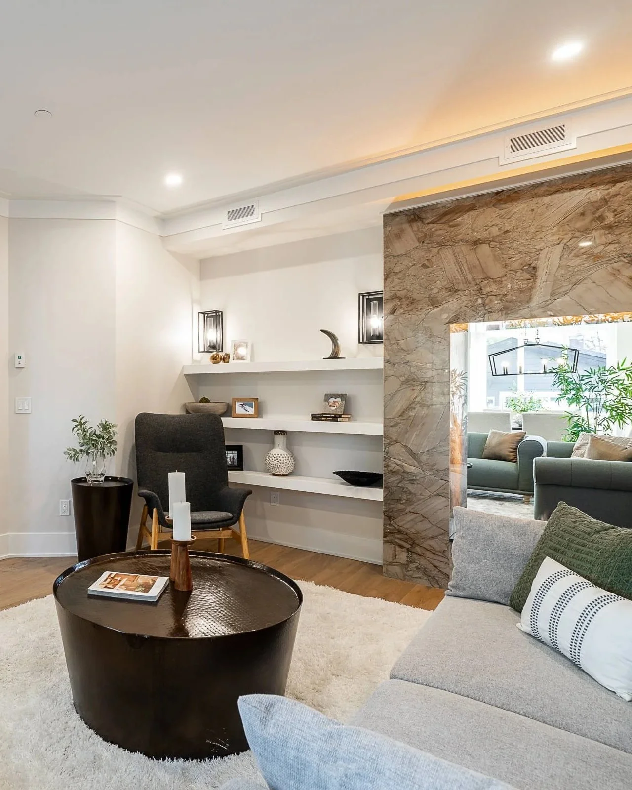 Modern living room with white walls, a black round coffee table with a candle and magazine, gray sofa with green and patterned pillows, a black armchair, white shelves with decor, a marble partition, and a view of another seating area with green sofa