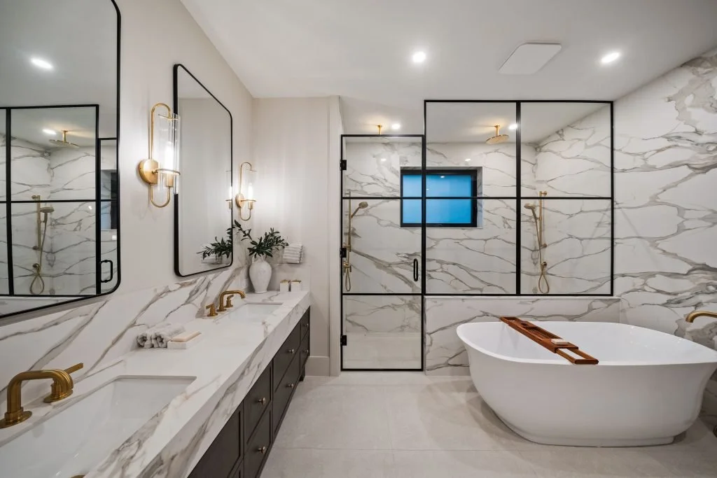 Modern bathroom with white marble walls and floor, black-framed mirrors and glass shower enclosure, freestanding bathtub, gold fixtures, and a double vanity with black cabinets and white marble countertop.
