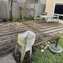 A backyard scene with wooden deck planks, a white plastic chair, a small table, and a patio umbrella base without an umbrella. There is a fence and some plants in the background.