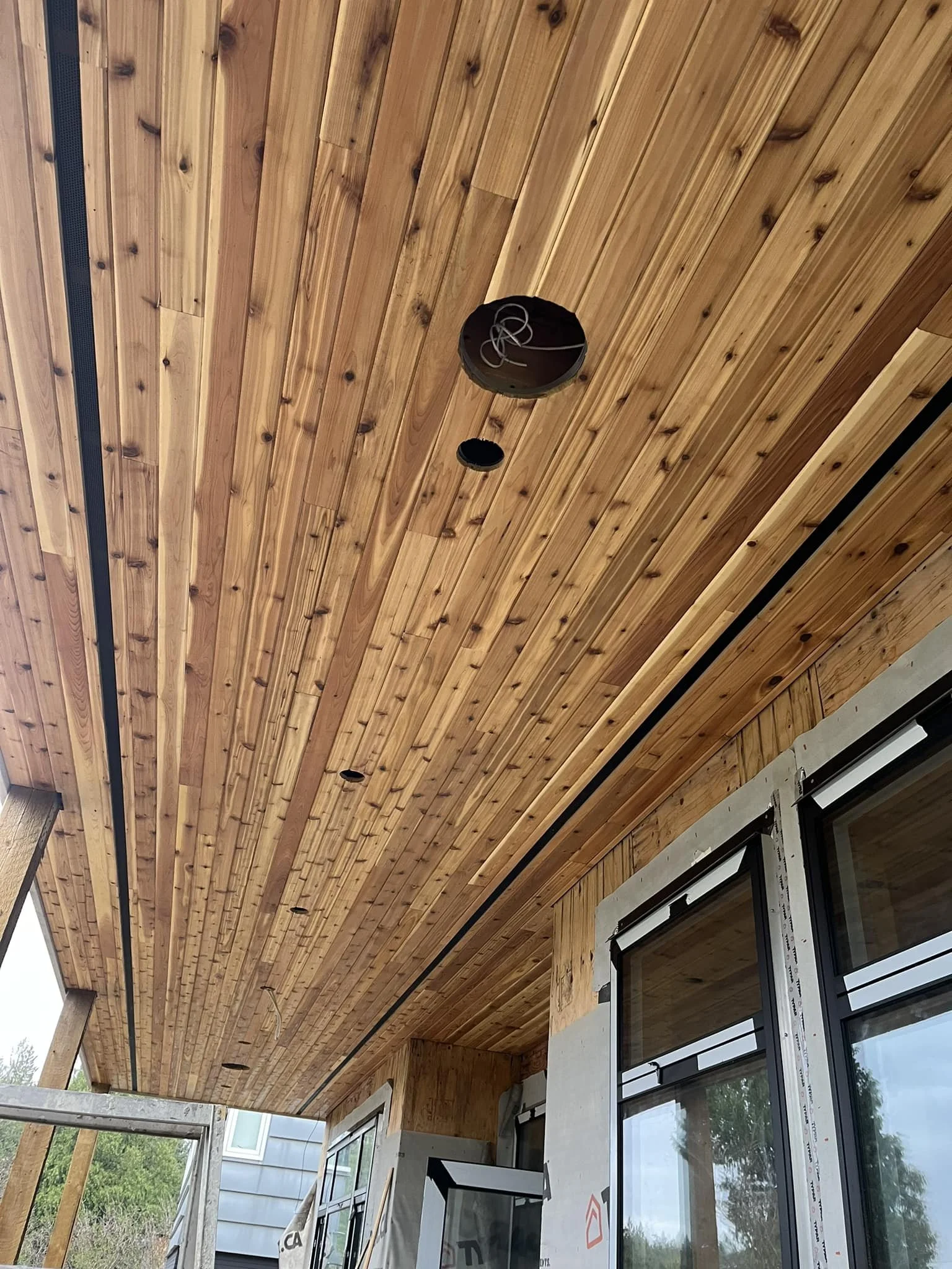 Wood-paneled ceiling with electrical wiring and openings for light fixtures, adjacent to a building exterior with windows and concrete wall