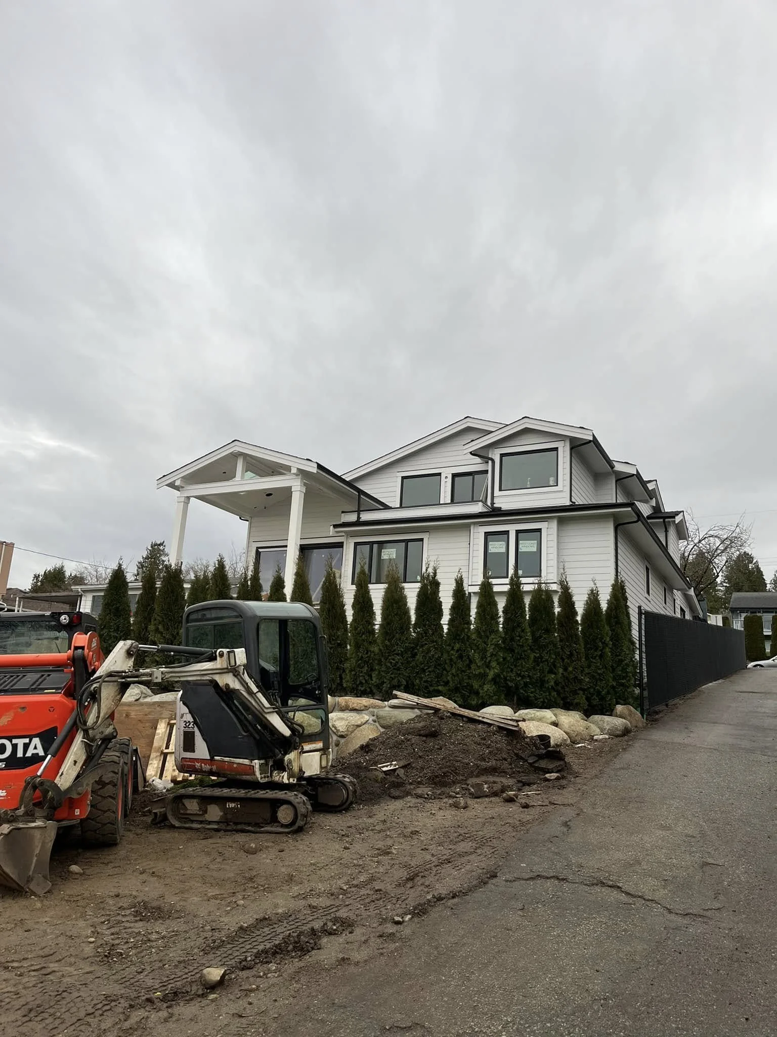Construction site with a new white multi-story house and two construction vehicles, one small excavator and one skid steer, on a dirt road under overcast sky.