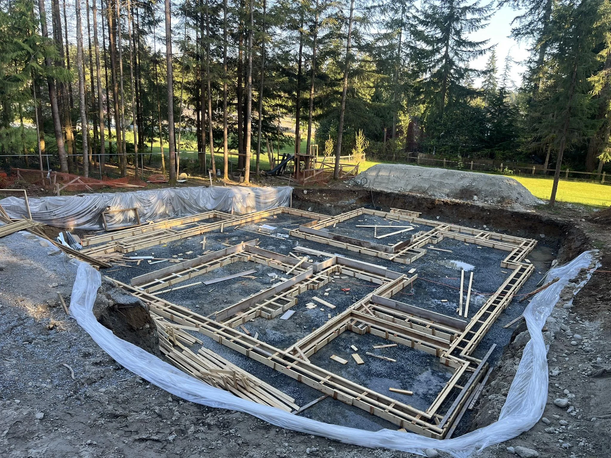 Construction site with wooden framing for a foundation, surrounded by plastic sheeting, in a wooded area with trees and grass.