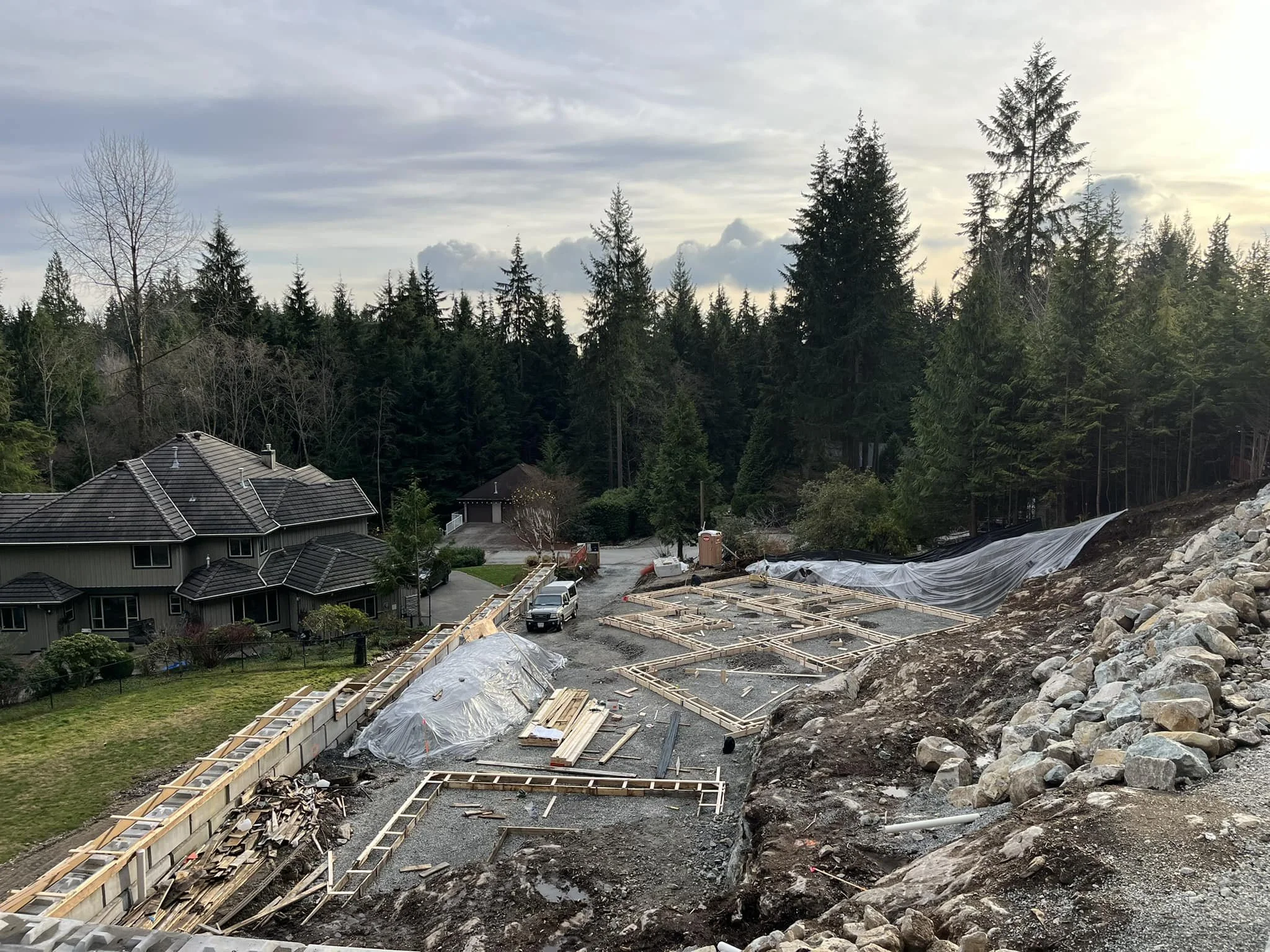 Construction site with wooden framework, gravel ground, and a partially built foundation under a cloudy sky, with a house and tall trees in the background.