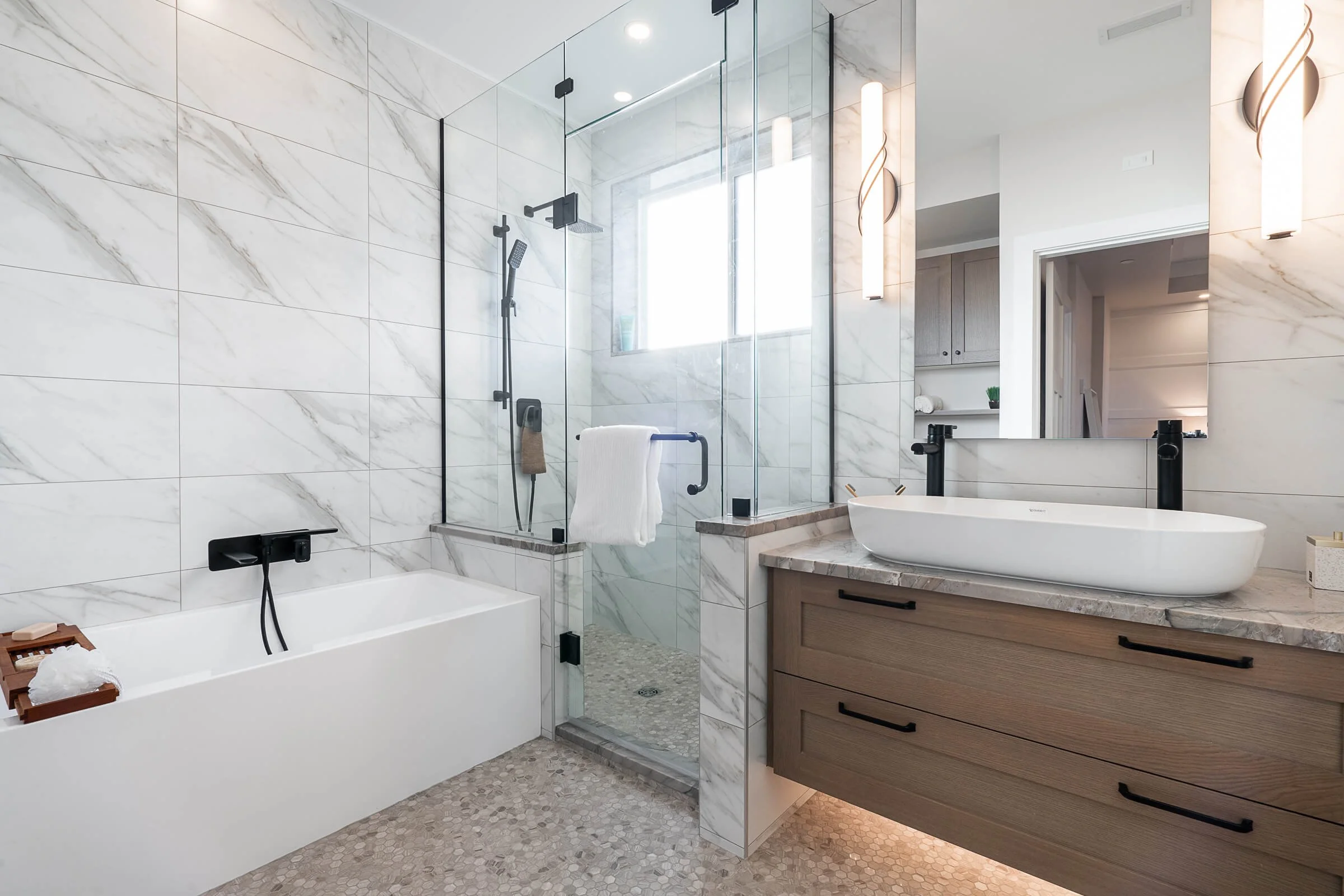 Modern bathroom with a bathtub, glass-enclosed shower, marble walls, wooden vanity with a vessel sink, large mirror, and contemporary lighting fixtures.
