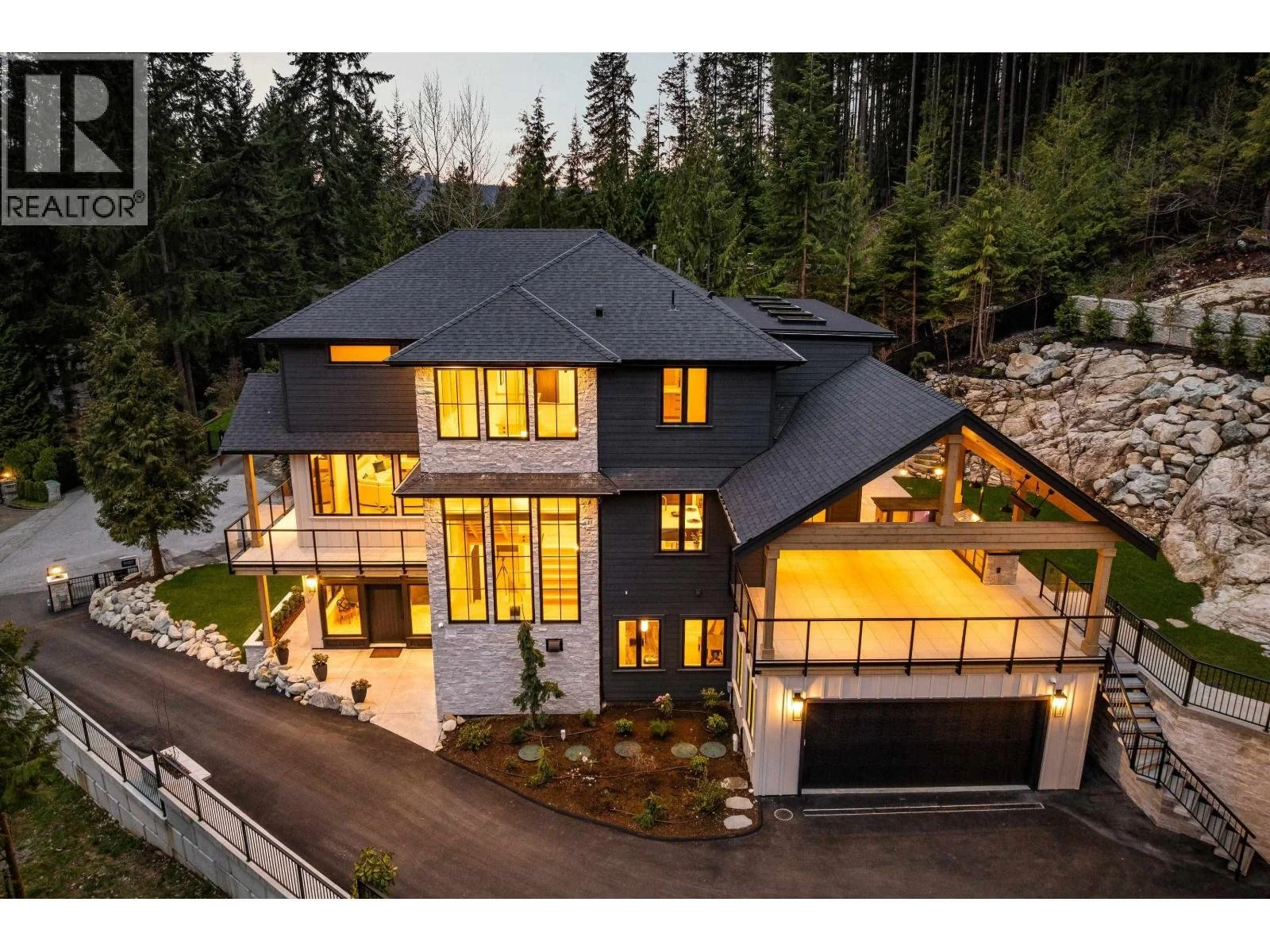 Large modern house with multiple levels, illuminated windows, black siding, stone accents, surrounded by trees and rocky landscape, with a driveway and a large deck area.