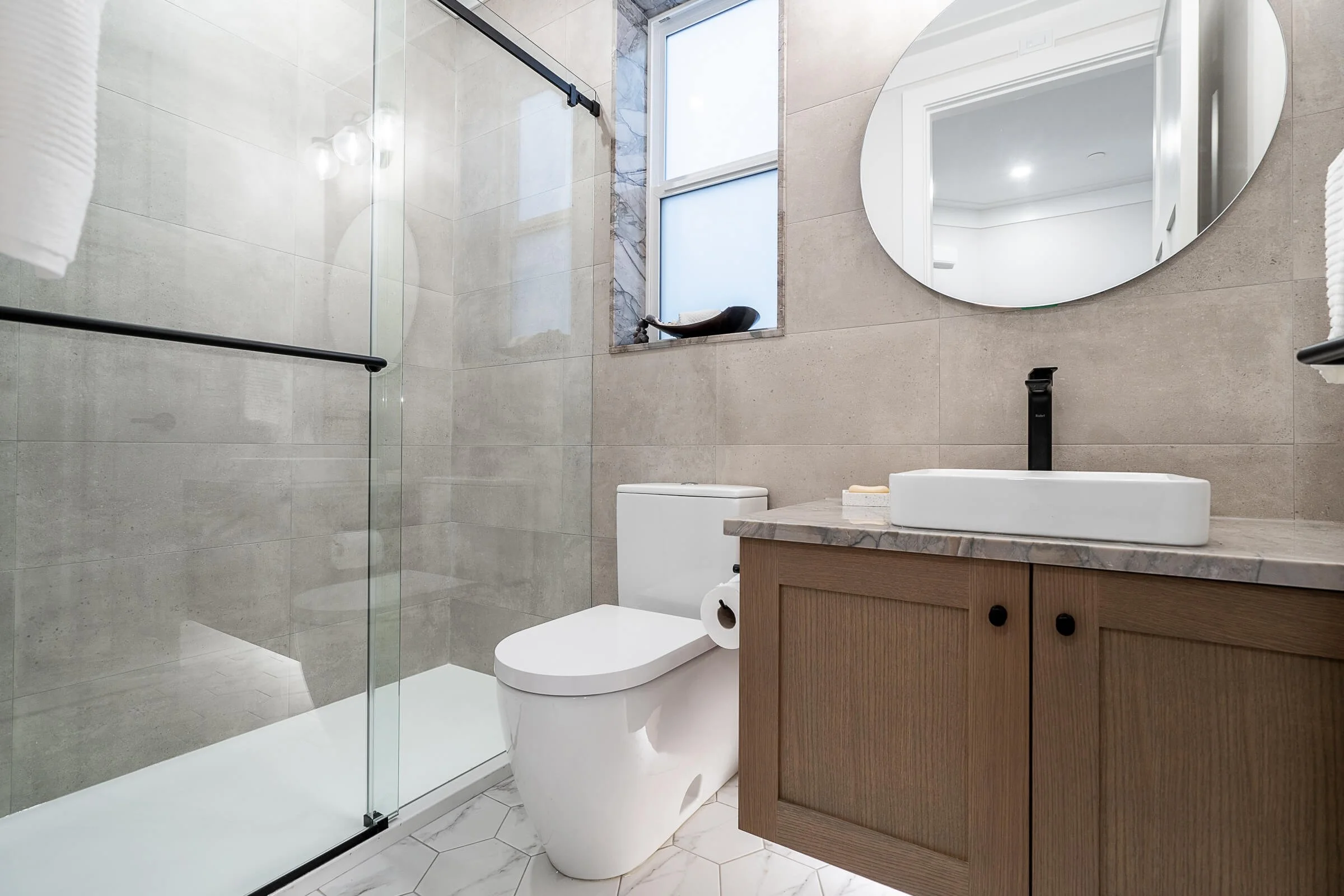 Modern bathroom with a glass shower enclosure, a white toilet, a wooden vanity with a marble countertop, and a round mirror above the sink, with a window providing natural light.
