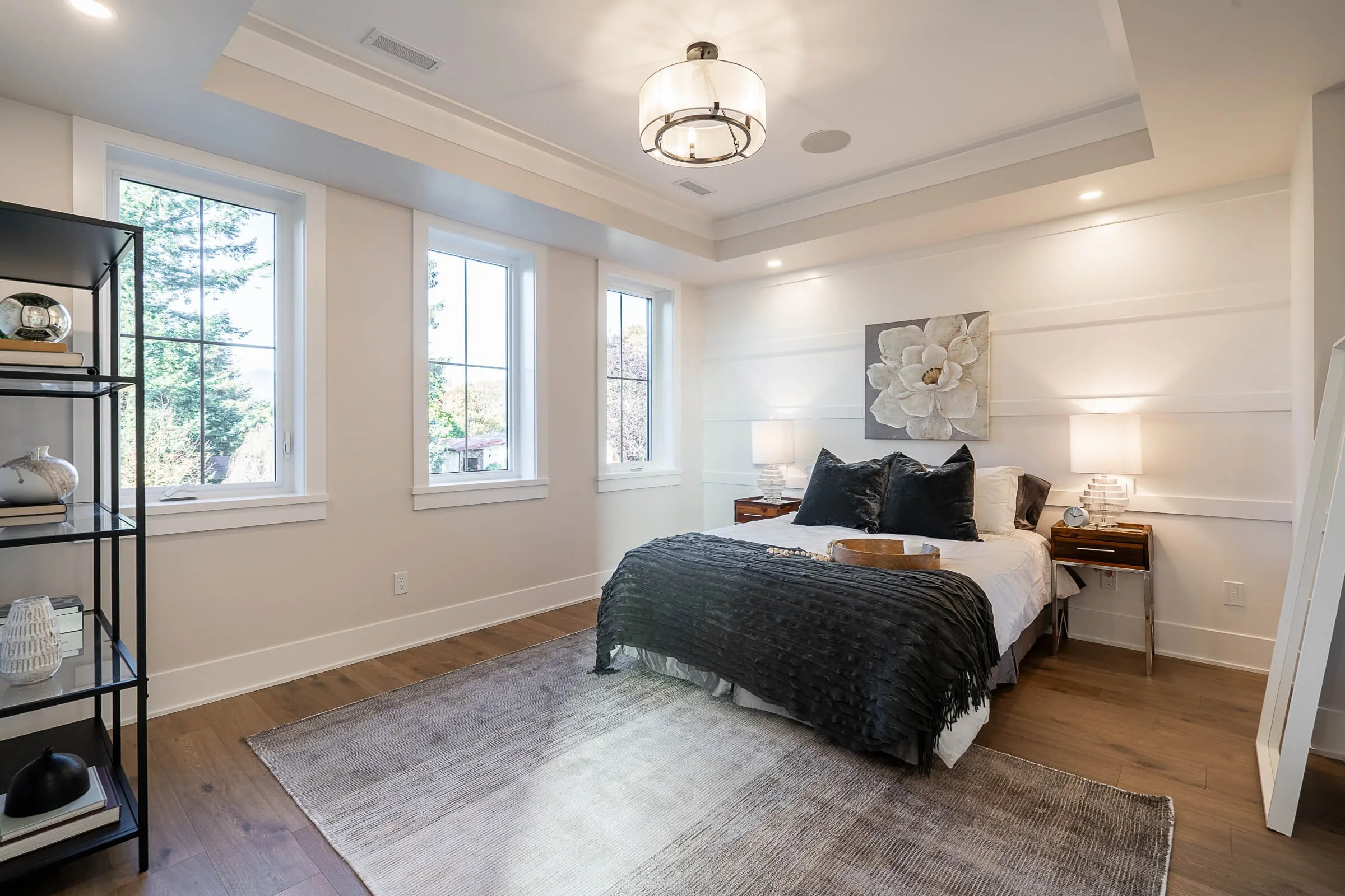 Bedroom with white walls, three windows, wooden floors, bed with black and white bedding, two nightstands with lamps, and a decorative flower painting above the bed.