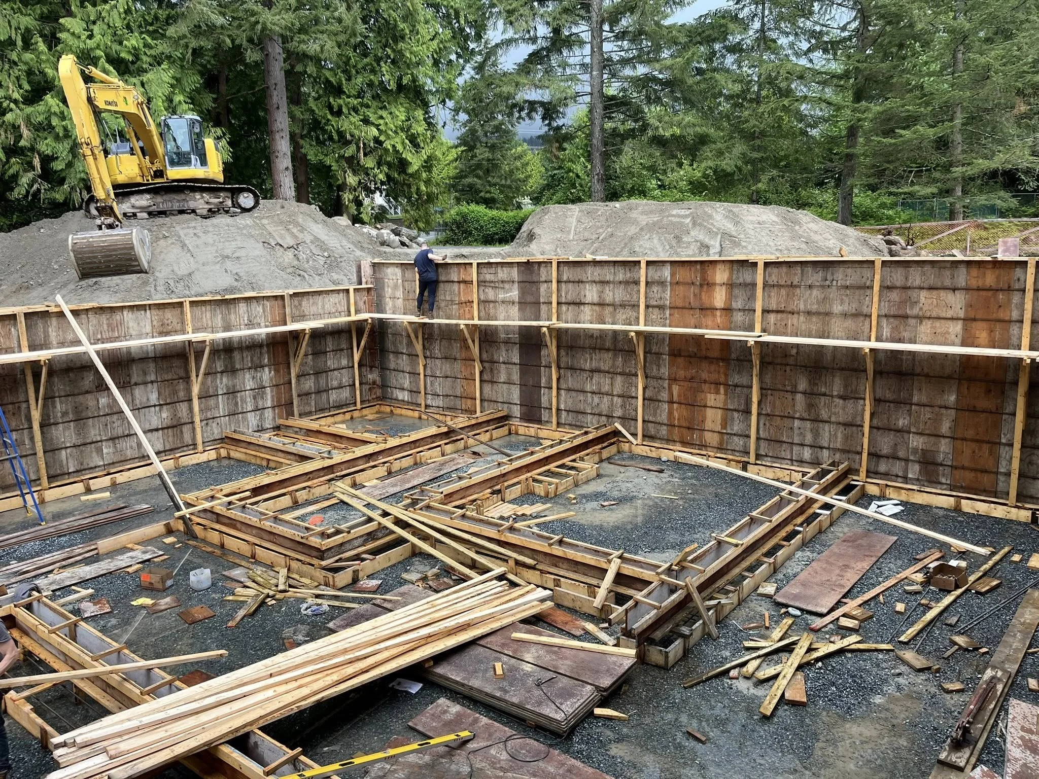 Construction site with wooden forms and scaffolding for building foundation, with a yellow excavator on a dirt mound and trees in the background.