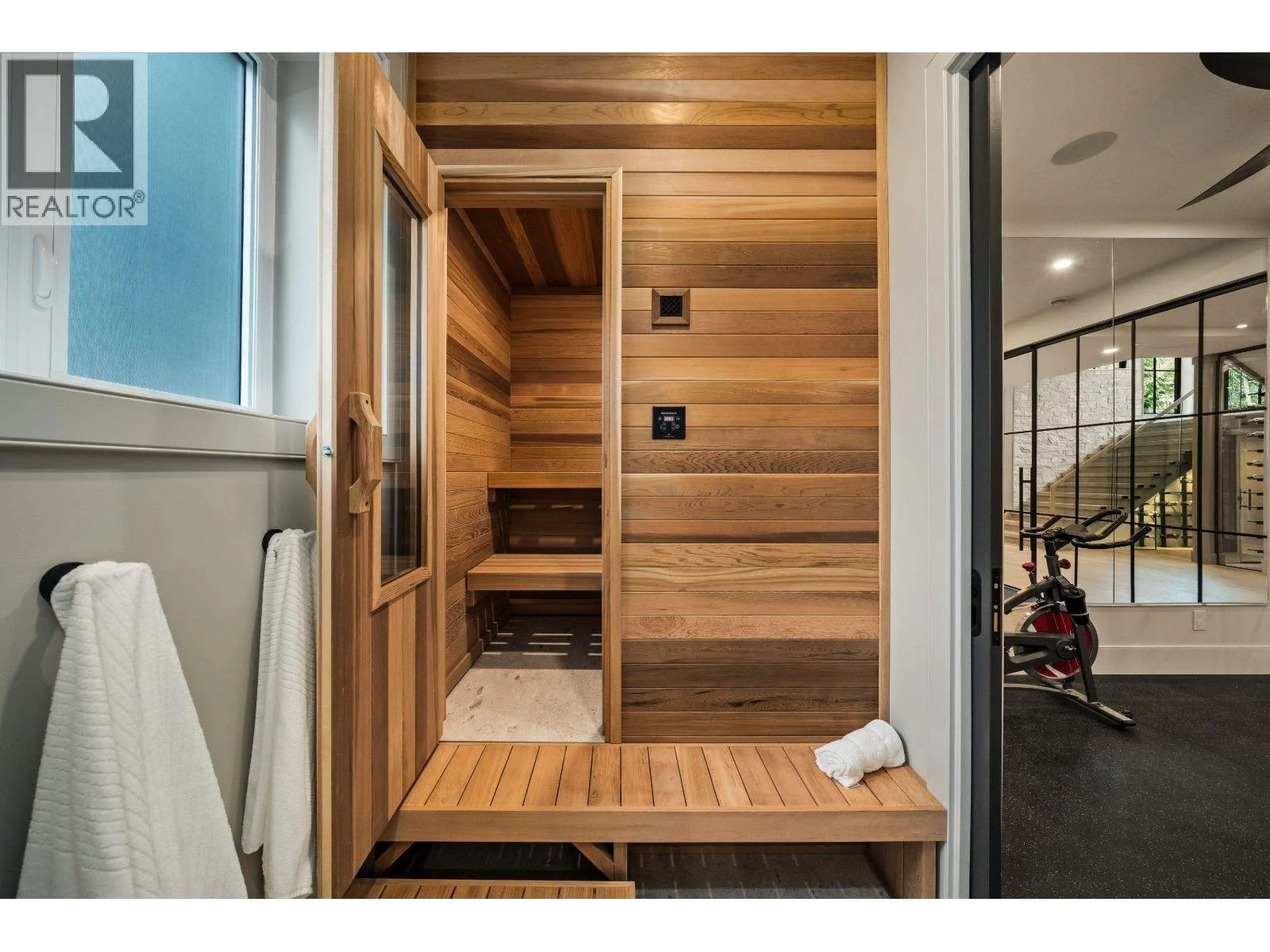 Indoor wood-paneled sauna with bench seating, located next to a window and a towel rack, adjacent to a fitness area with exercise bike and stairs.