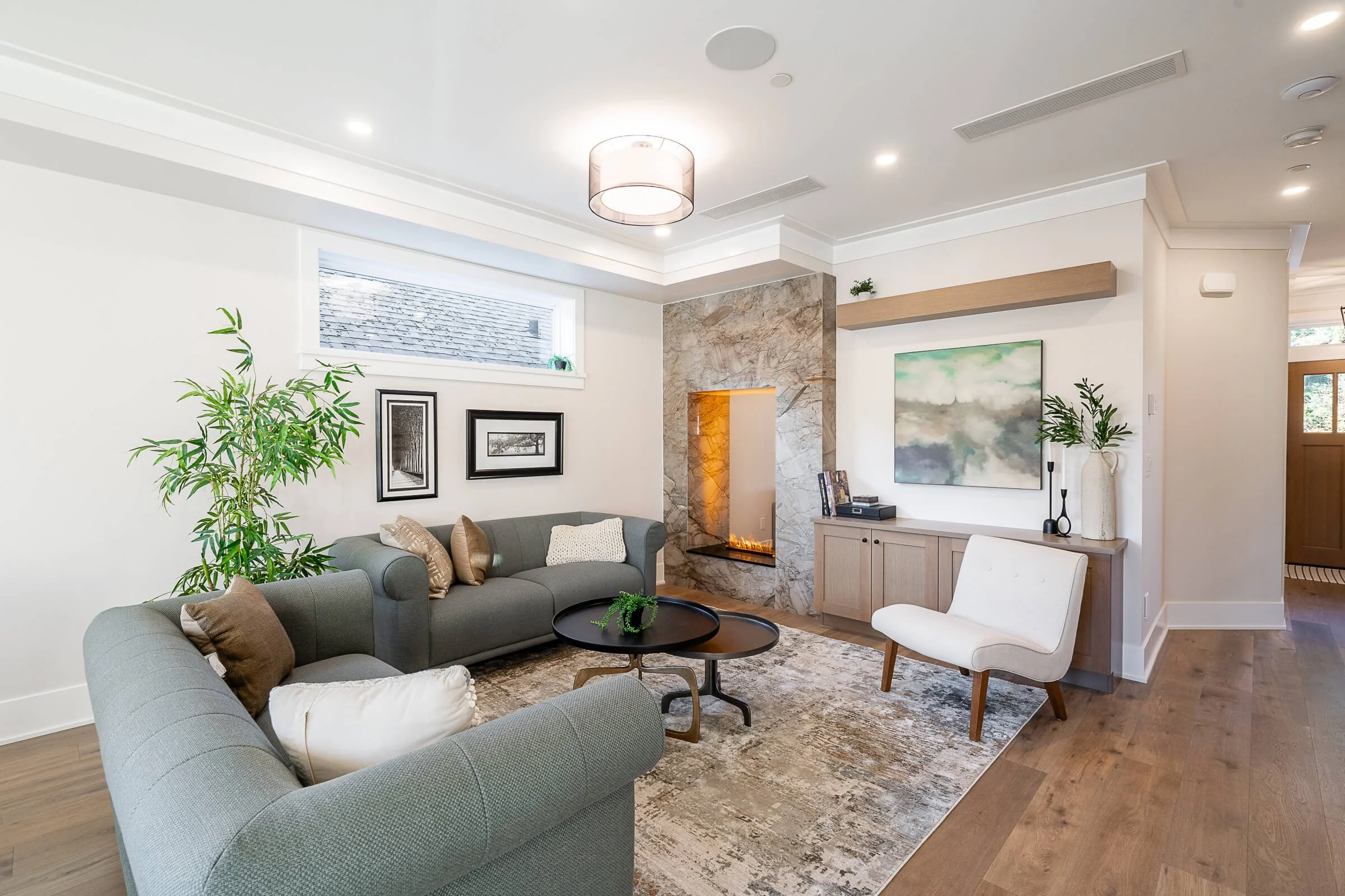 Modern living room with a gray sofa, black coffee table, white and brown pillows, a tall green plant, beige and gray area rug, a fireplace with marble surround, abstract art on the wall, a white armchair, and wooden flooring.