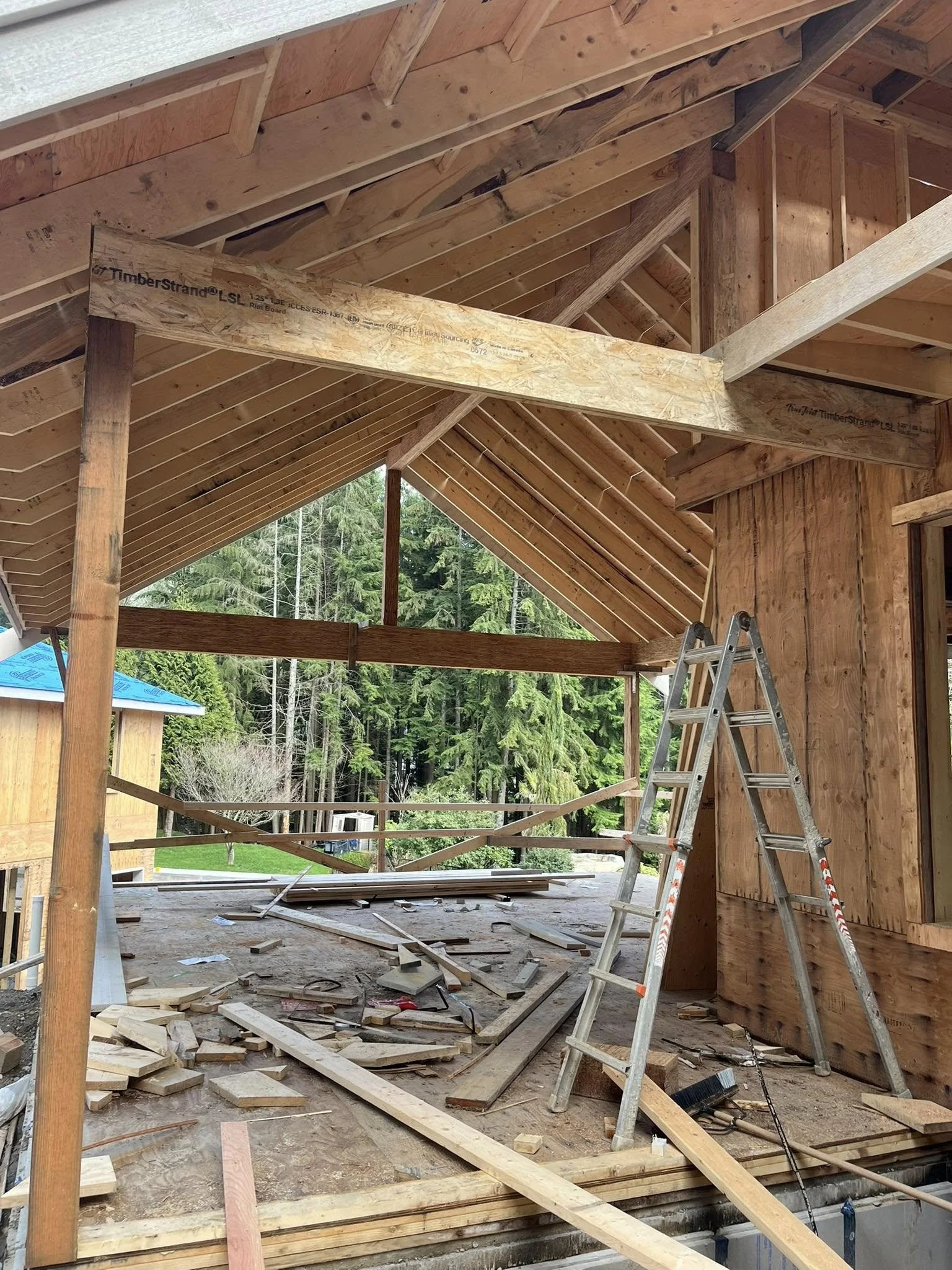 Construction site inside a wooden house with exposed framing, an aluminum ladder, and scattered construction materials, overlooking a background of green trees.