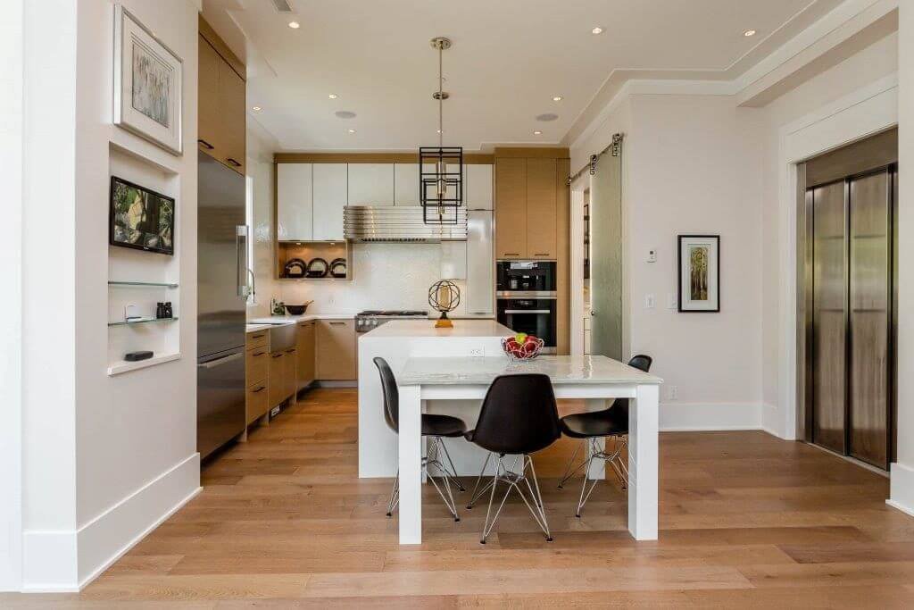 Modern kitchen with white and tan cabinets, a white island with black chairs, stainless steel refrigerator, oven, and sliding glass door, hardwood flooring, and decorative lighting.