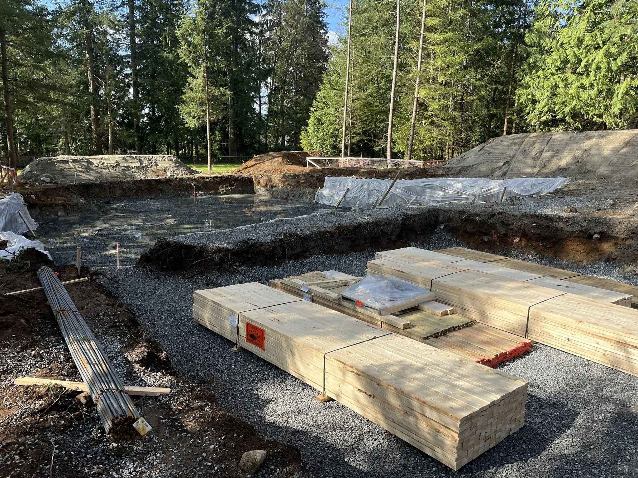 Construction site with large stacks of wooden planks, metal pipes, and wrapped materials, surrounded by dirt and gravel, with trees and blue sky in the background.